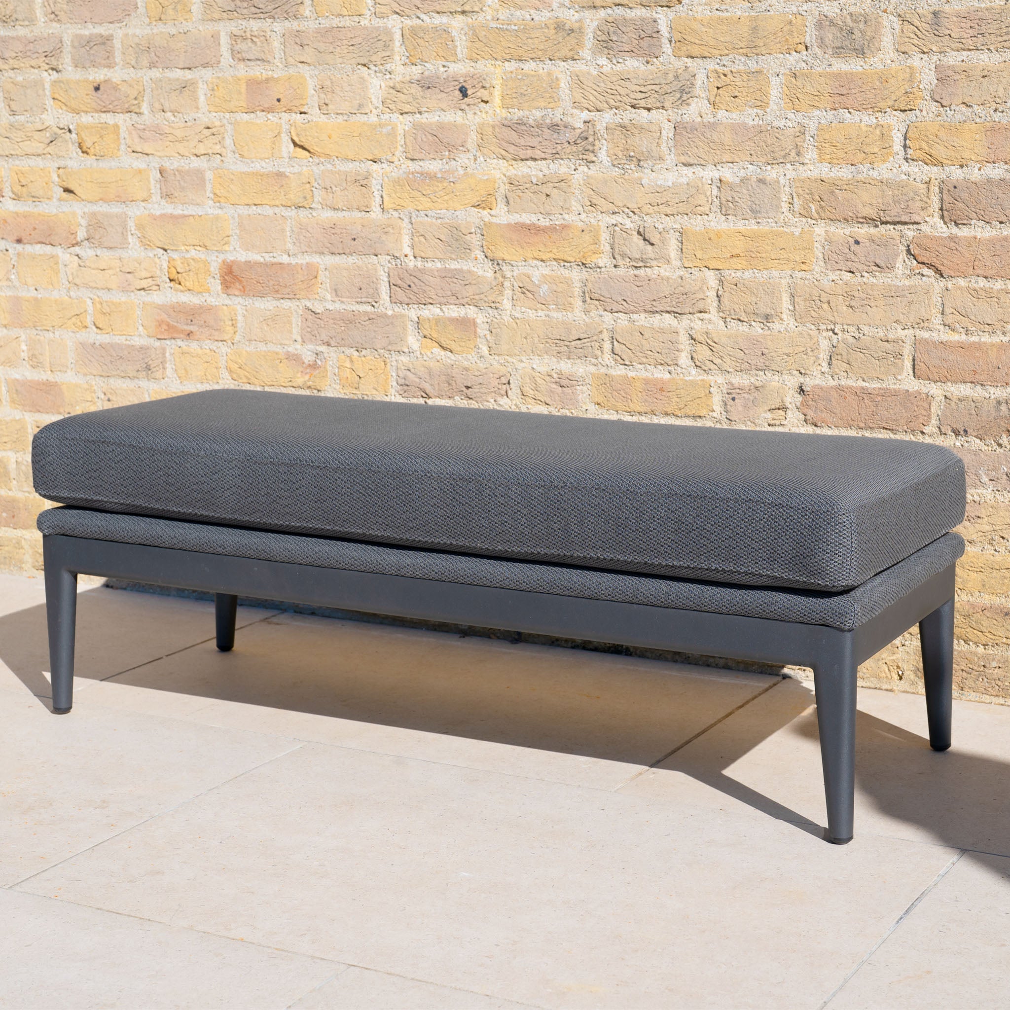 The Luna Bench in Grey, featuring weather-resistant upholstery and black legs, provides stylish outdoor seating on a light stone patio beside a tan brick wall.