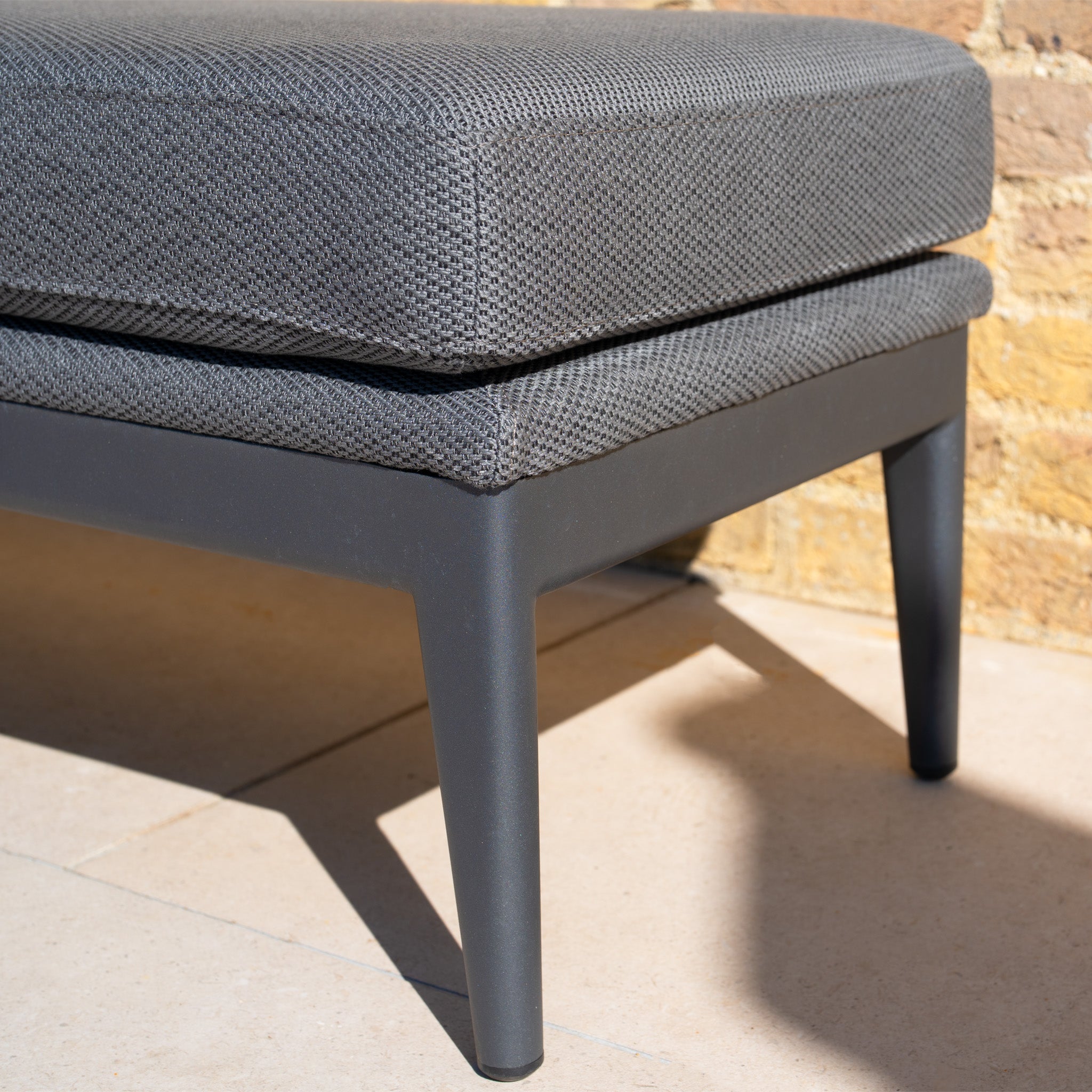 Close-up of a modern outdoor bench with a textured gray cushion and black metal legs on a tiled floor, paired with the stylish Luna Stool in Grey for a chic outdoor furniture setup.