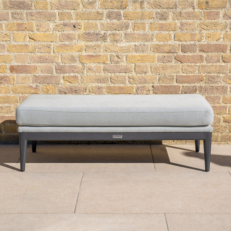 The Luna Bench in Oyster Grey features a light gray upholstered design with dark legs, ideal for stylish outdoor lounging and all-weather durability on your patio.