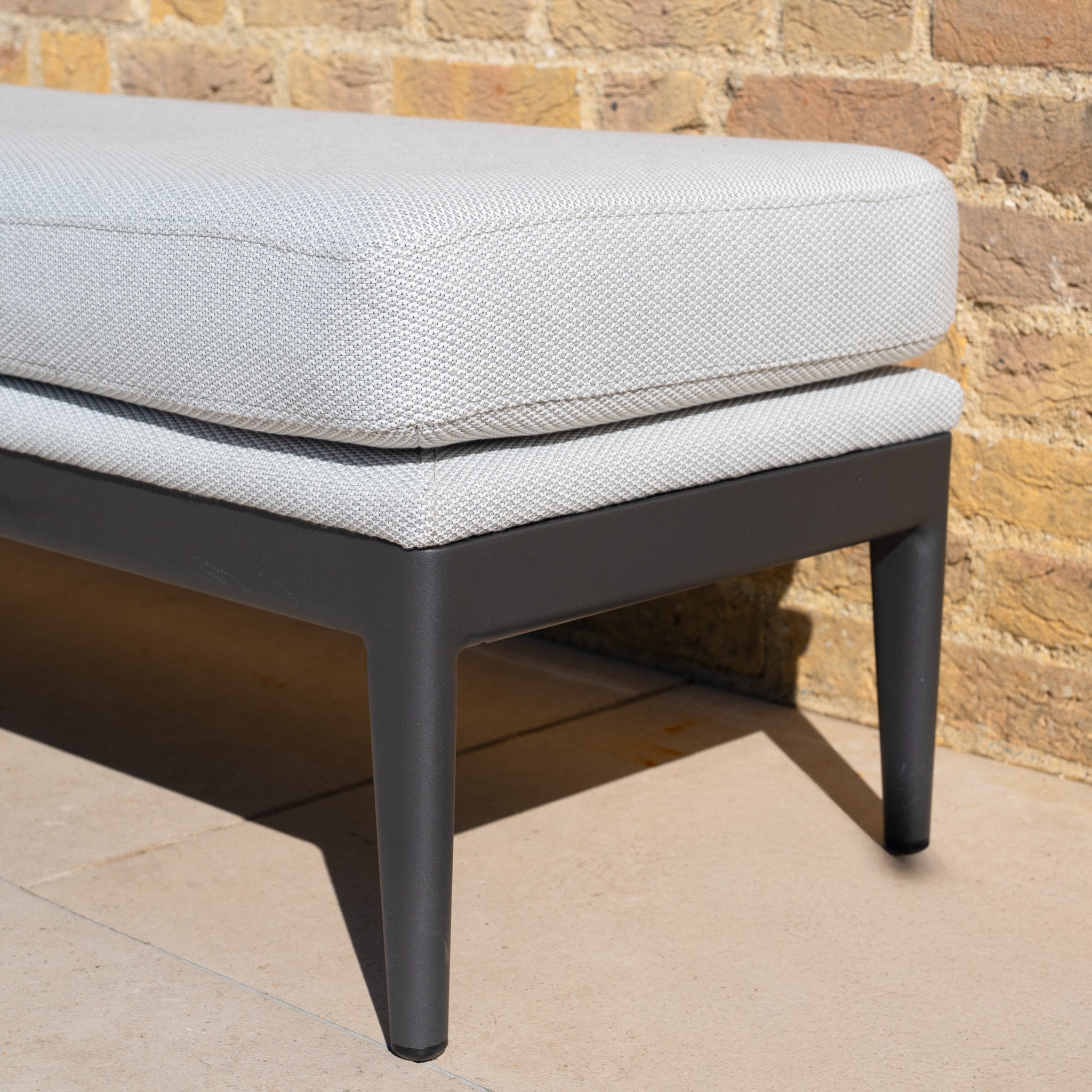 Close-up of the Luna Bench in Oyster Grey, an outdoor bench with a light gray cushion and dark metal legs, set against a brick wall.