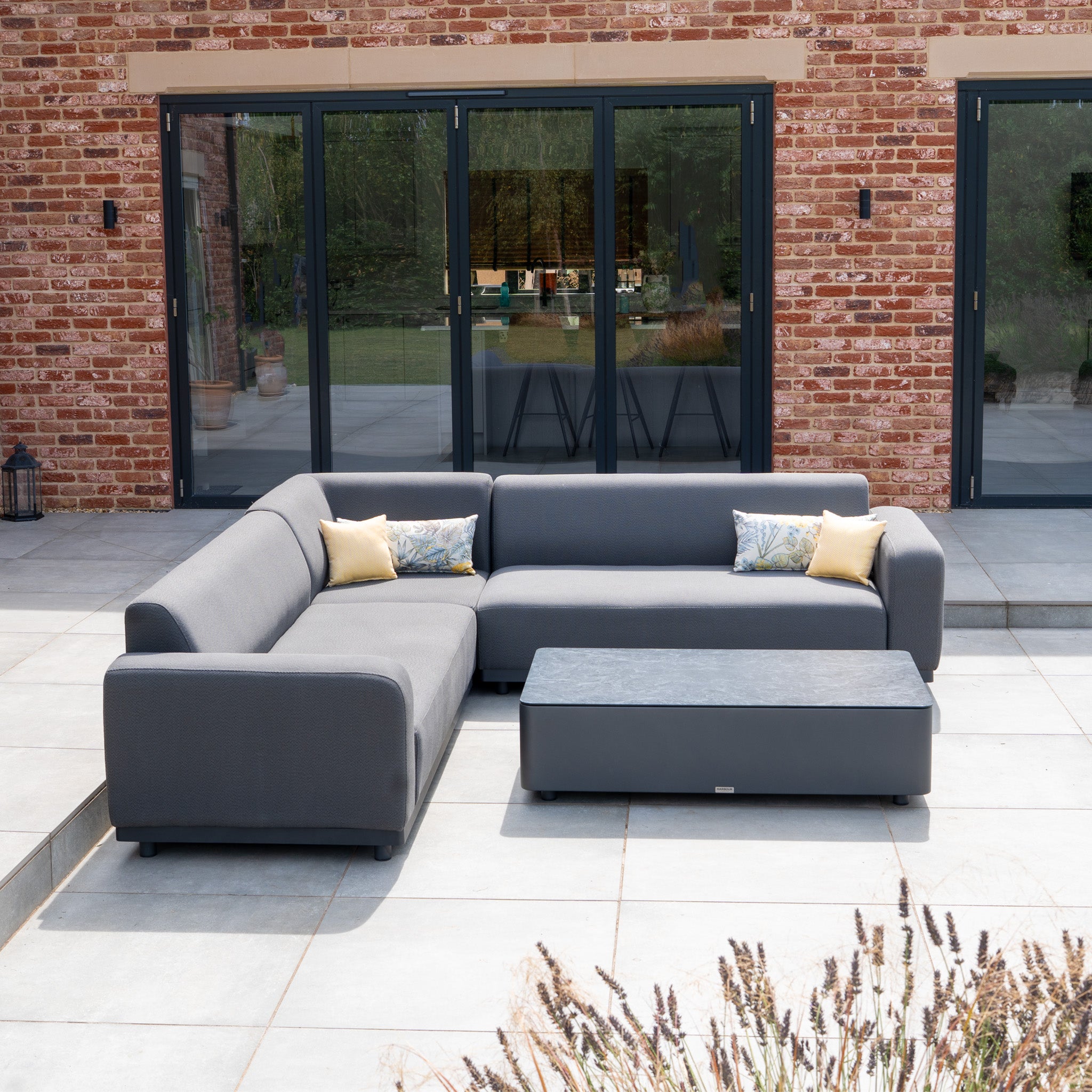 The Luna Outdoor Fabric Corner Sofa Set with Coffee Table in Grey features stain-proof cushions, perfect for patios by glass doors and brick walls.