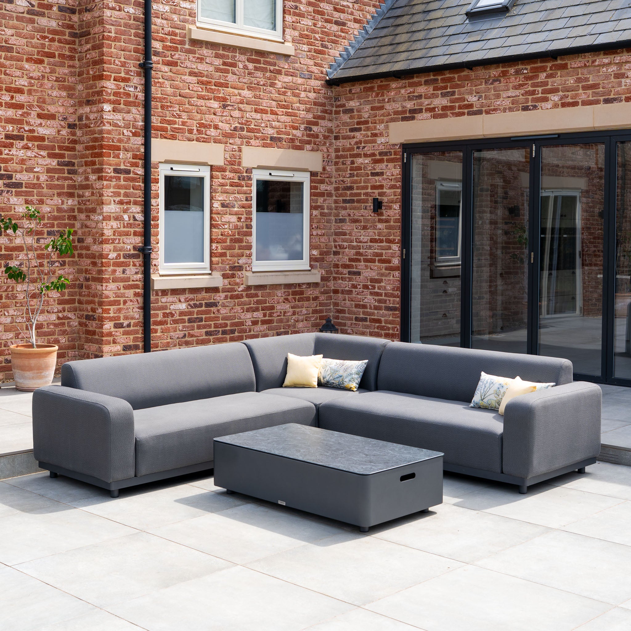 The Luna Outdoor Fabric Corner Sofa Set with Coffee Table in Grey features durable outdoor cushions and a sleek table, ideal for patios next to a brick house.