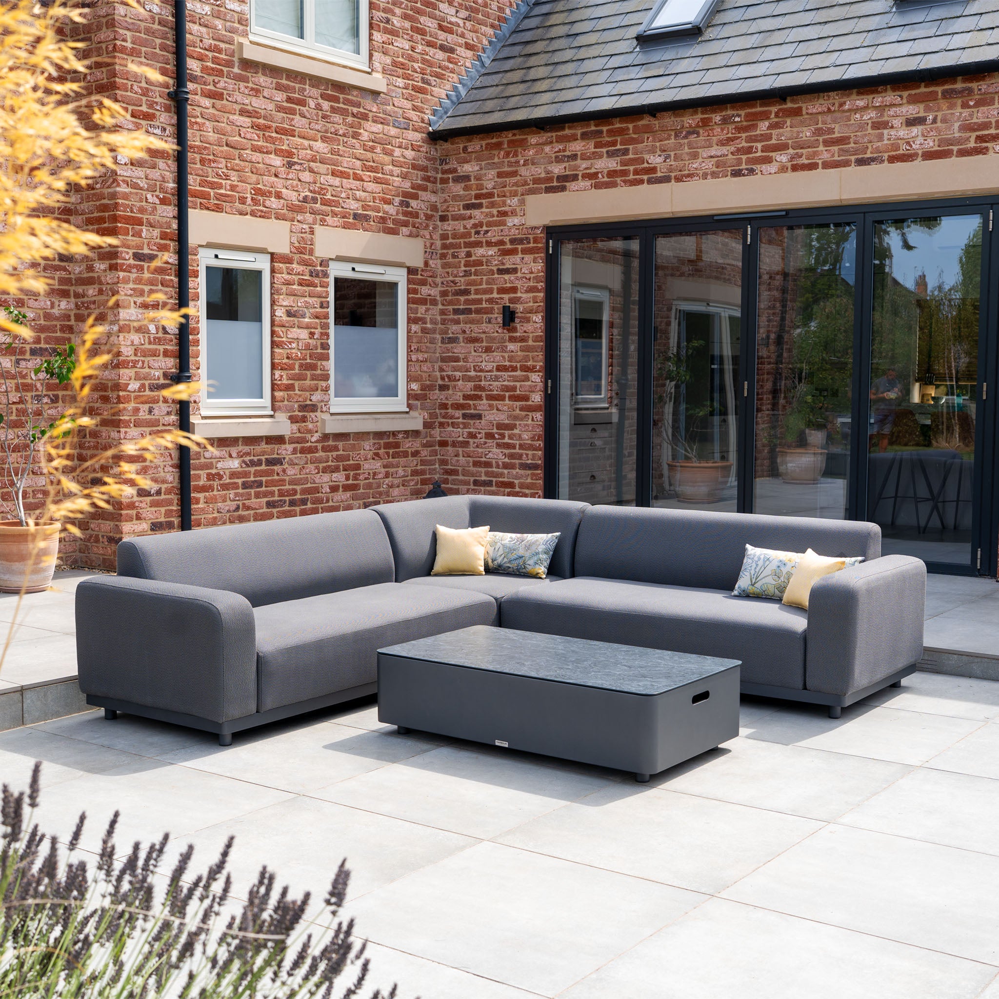 The Luna Outdoor Fabric Corner Sofa Set with Coffee Table in Grey features stain-proof cushions and modern design, perfect for patios beside a brick house.
