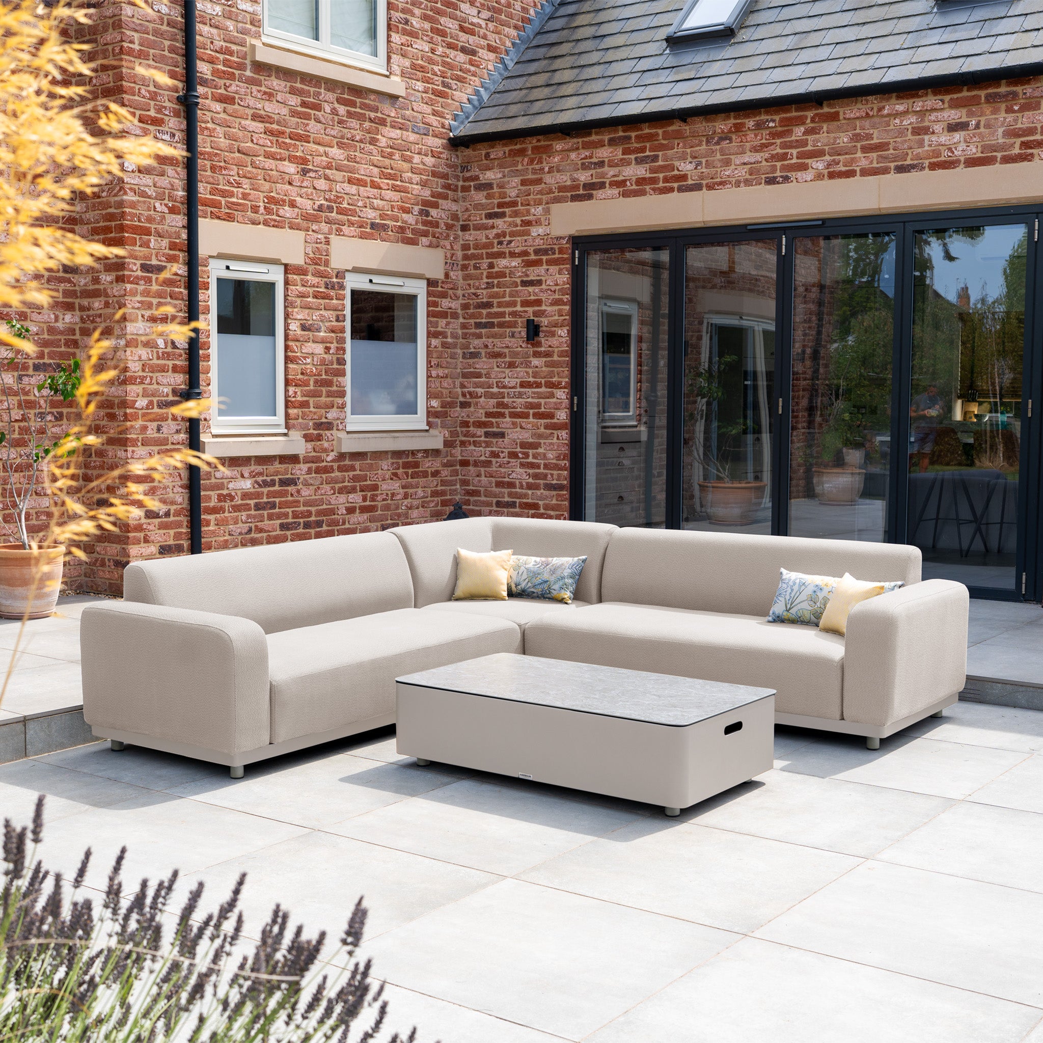 The Luna Outdoor Fabric Corner Sofa Set with Coffee Table in Fawn sits on a patio by a brick house with large windows, offering stain-proof, modern garden furniture for stylish and low-maintenance lounging.