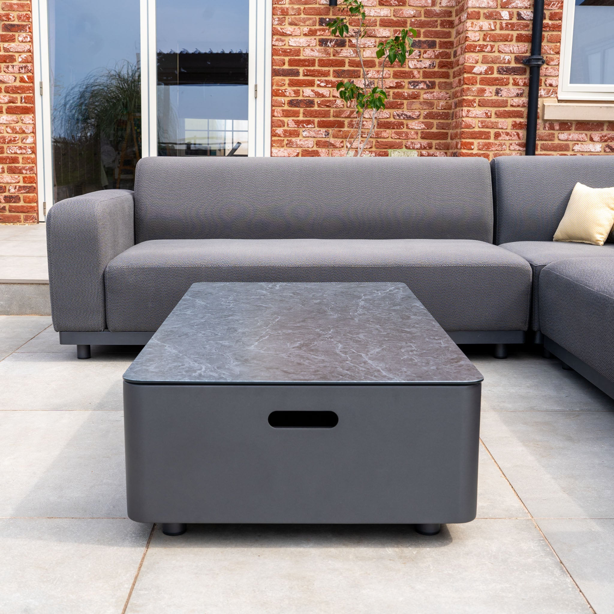 The Luna Outdoor Fabric Corner Sofa Set with Coffee Table in Grey features stain-proof upholstery, a sleek rectangular black table, and modern design—perfect for any tiled patio in front of a brick wall.