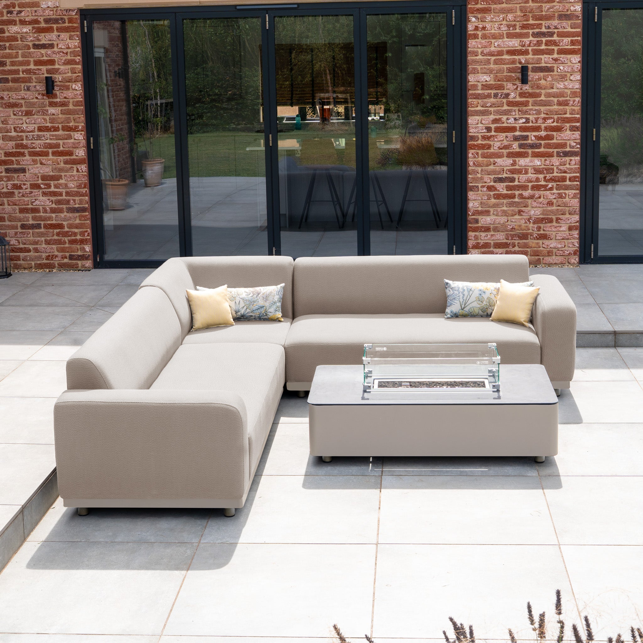 The Luna Outdoor Fabric Corner Sofa Set with Firepit Coffee Table in Fawn—stylish, all-weather patio furniture perfect for your backyard retreat, placed in front of large glass doors.