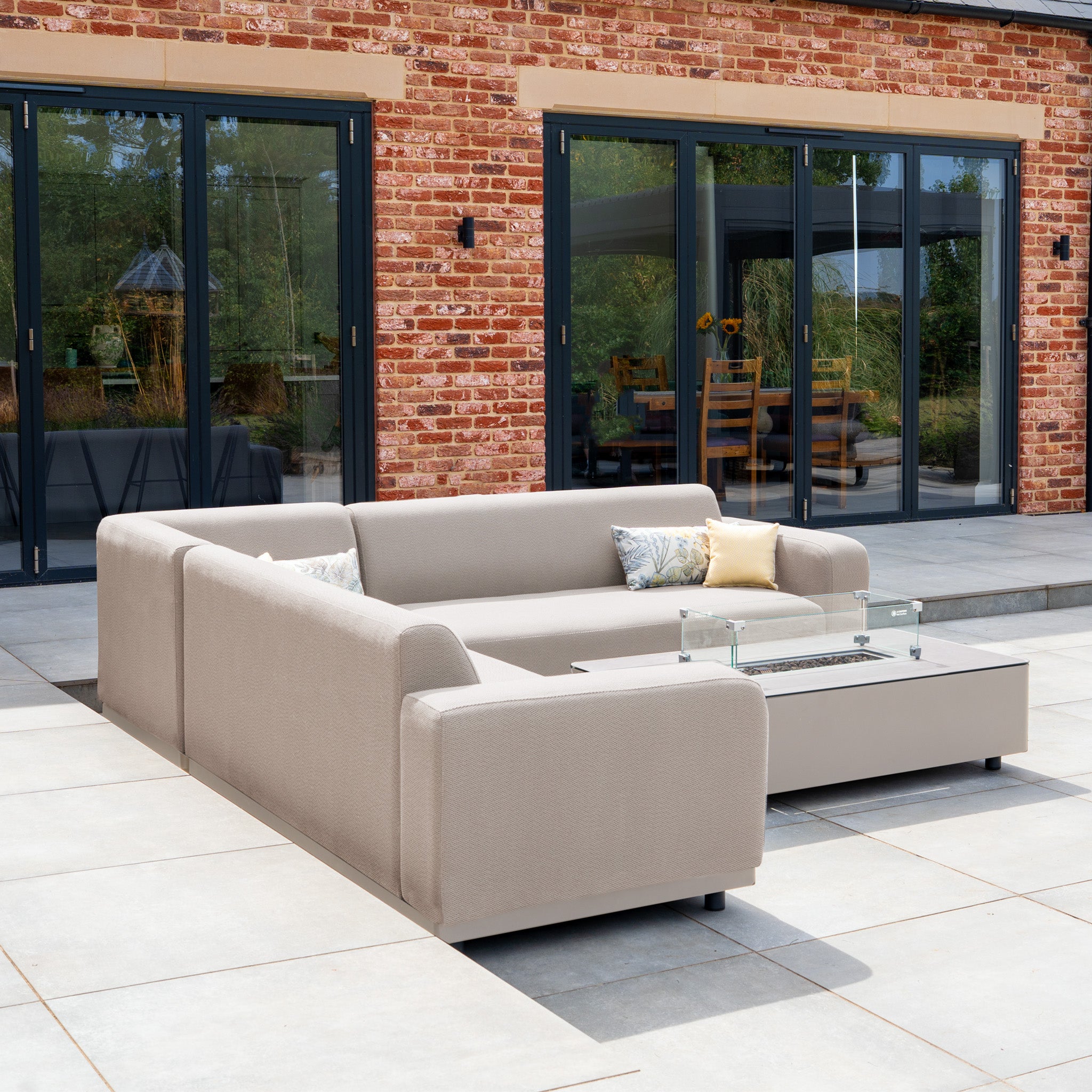 The Luna Outdoor Fabric Corner Sofa Set with Firepit Coffee Table in Fawn is arranged on a patio before glass doors, featuring plush cushions and a cozy firepit centerpiece.
