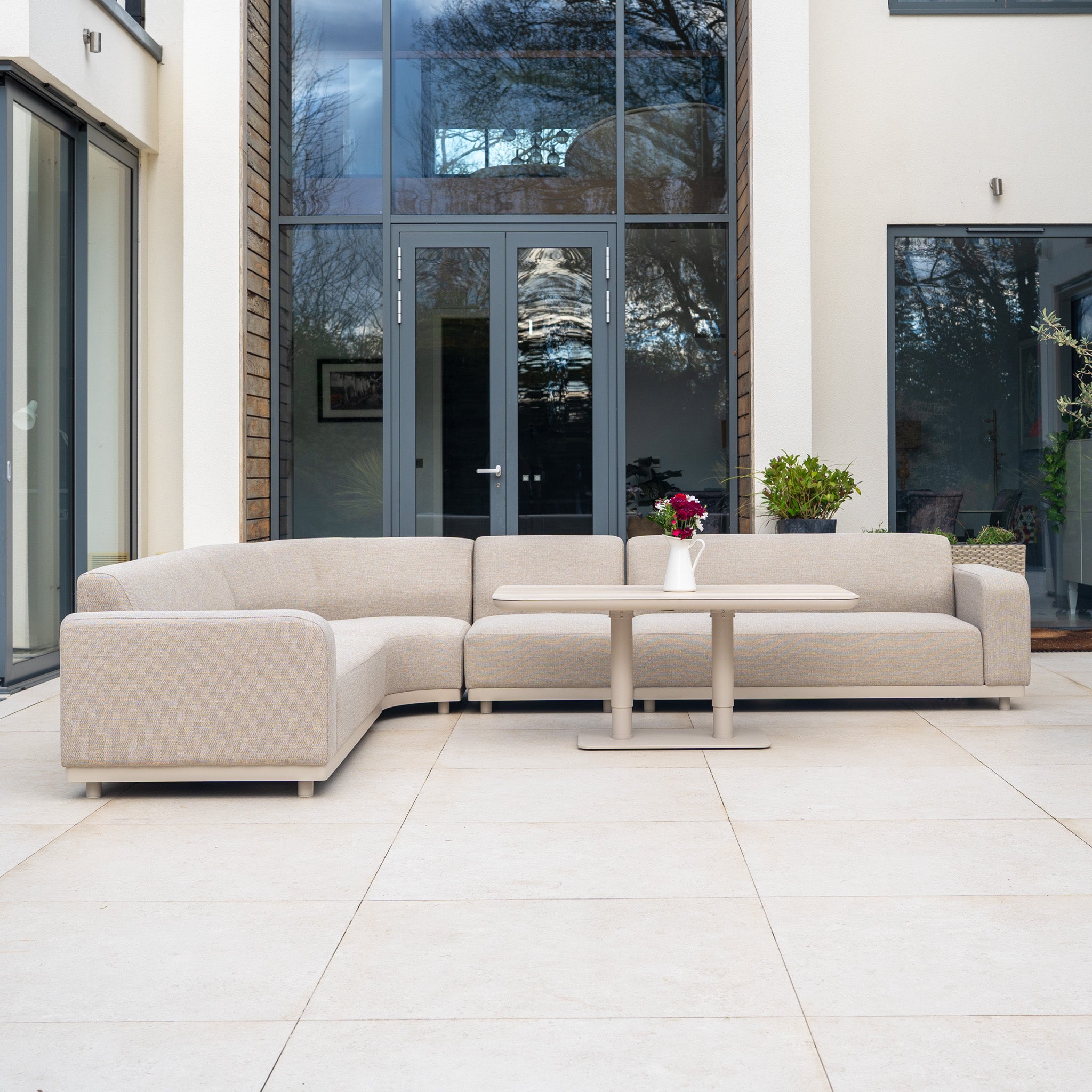 The Luna Outdoor Fabric Large Curved Corner Sofa Set with Altura Rising Table in Fawn sits on a tiled patio of a modern home, featuring quick-dry cushions and plants with flowers arranged on the table.