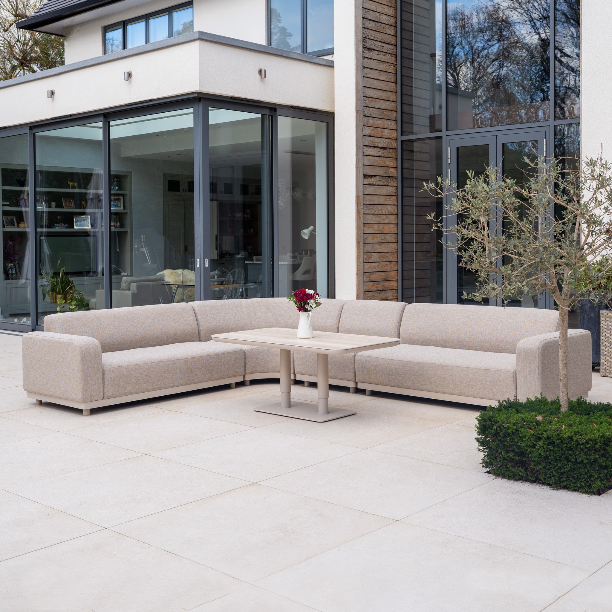 Modern patio featuring the Luna Outdoor Fabric Large Curved Corner Sofa Set in Fawn with quick-dry cushions and an Altura Rising Table, accented with flowers and a contemporary glass house in the background.
