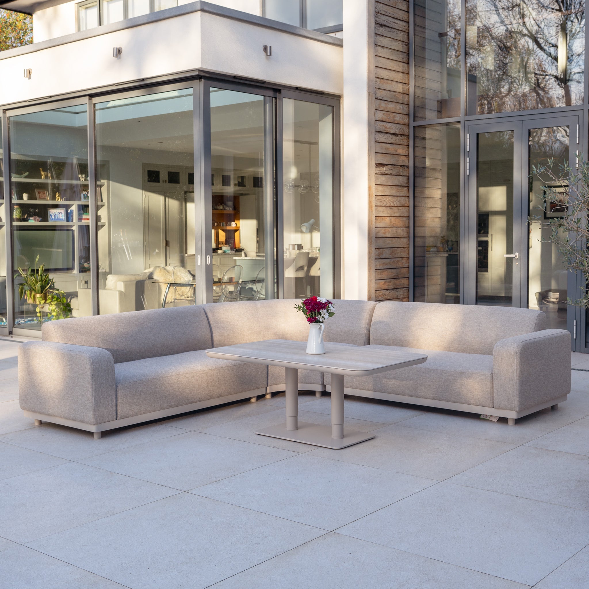 The Luna Outdoor Fabric Curved Corner Sofa Set with Altura Rising Table in Fawn creates an elegant modern patio scene outside a glass-walled house, showcasing stylish all-weather garden furniture.