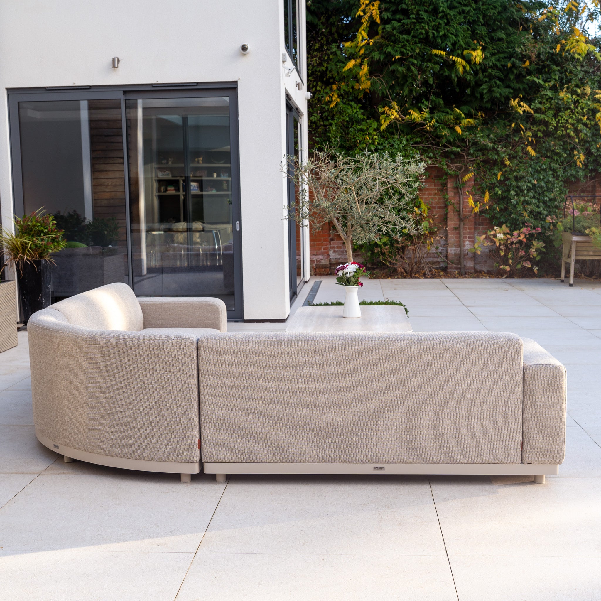 The Luna Outdoor Fabric Large Curved Corner Sofa Set with Altura Rising Table in Fawn features quick-dry cushions on a patio near a white house, complemented by plants and a small white table with flowers.