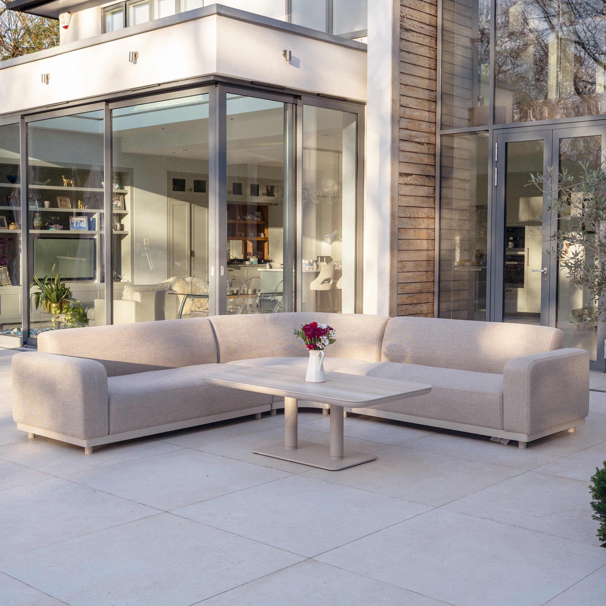 Modern patio featuring the Luna Outdoor Fabric Curved Corner Sofa Set with Altura Rising Table in Fawn, arranged by expansive glass doors and topped with a flower vase.