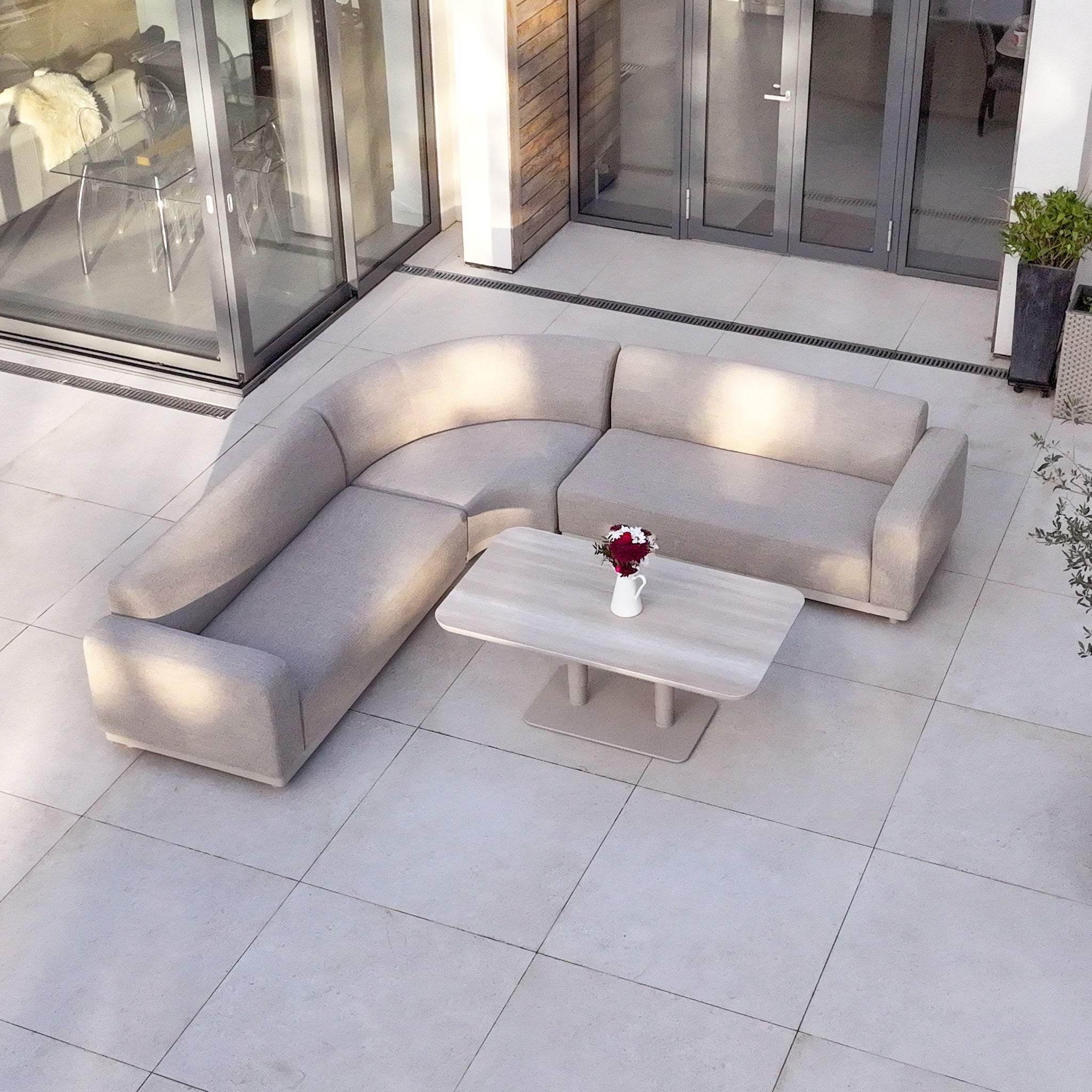 Modern outdoor patio featuring the Luna Outdoor Fabric Curved Corner Sofa Set with Altura Rising Table in Fawn, topped with a vase of red and white flowers.