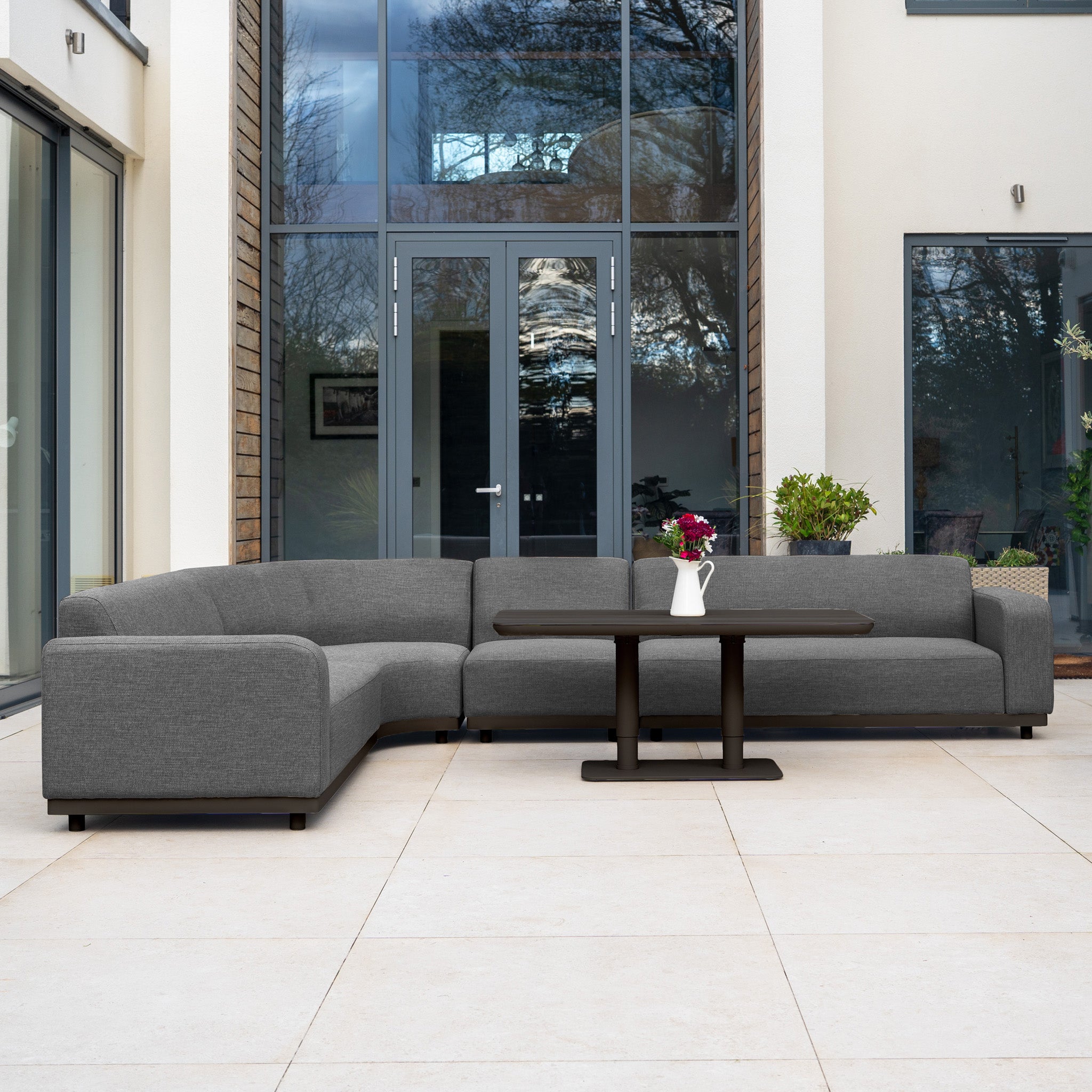 The Luna Outdoor Fabric Large Curved Corner Sofa Set with Altura Rising Table in Grey adds comfort and style to any modern patio, featuring durable all-weather fabric, elegant curves, and a versatile dark table.