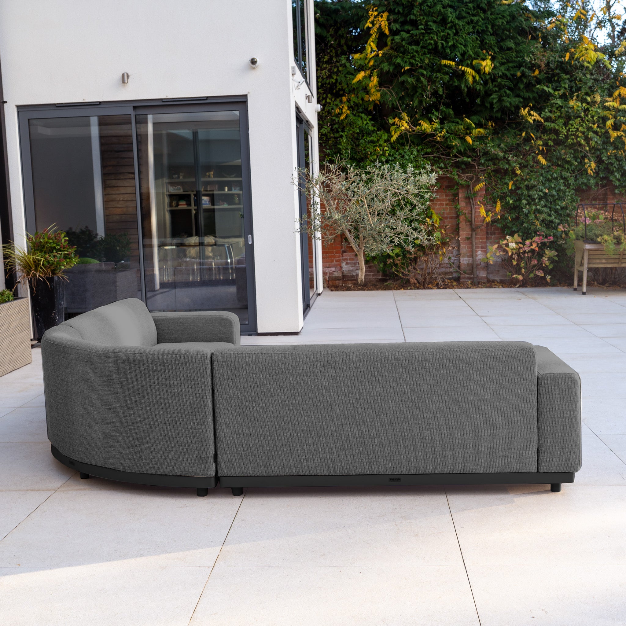 The Luna Outdoor Fabric Curved Corner Sofa Set with Altura Rising Table in Grey offers all-weather comfort and style, perfect for spacious patios with plants and large glass doors.