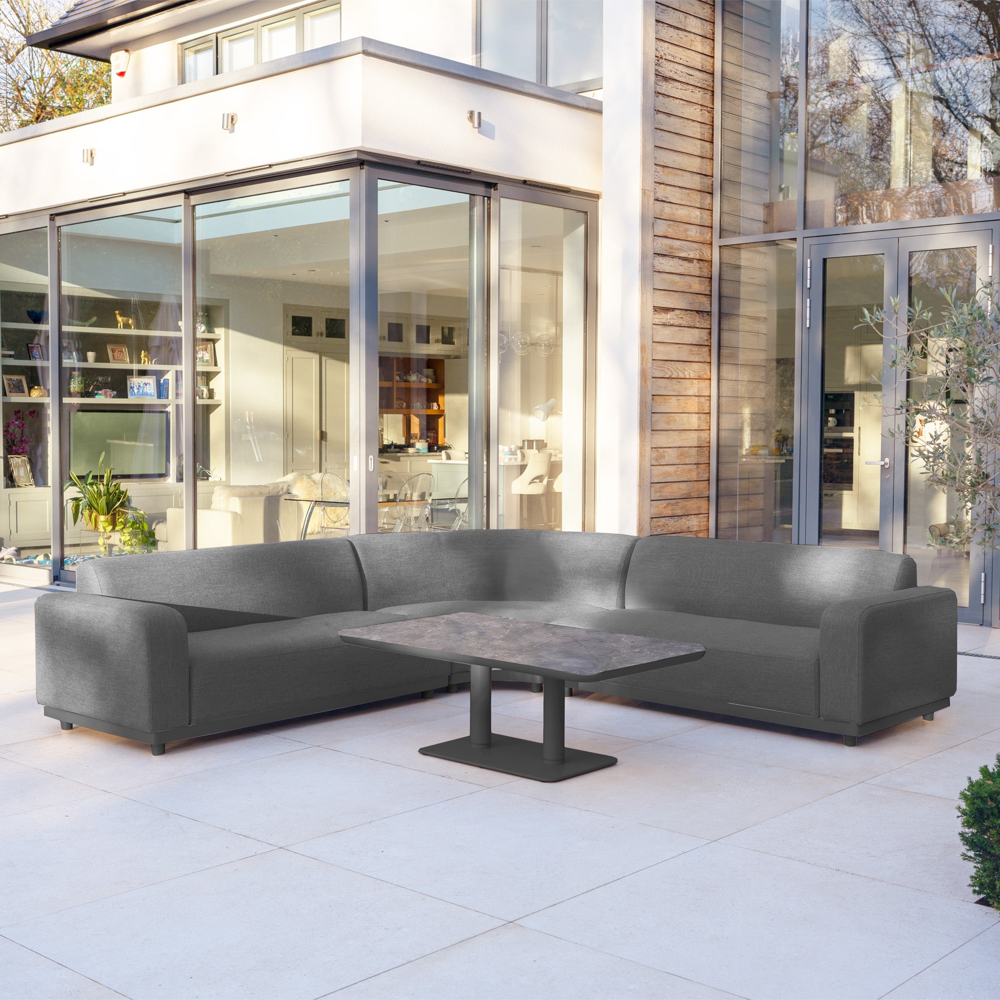 Modern patio featuring the Luna Outdoor Fabric Curved Corner Sofa Set with Altura Rising Table in Grey, positioned in front of glass doors and a wooden wall.