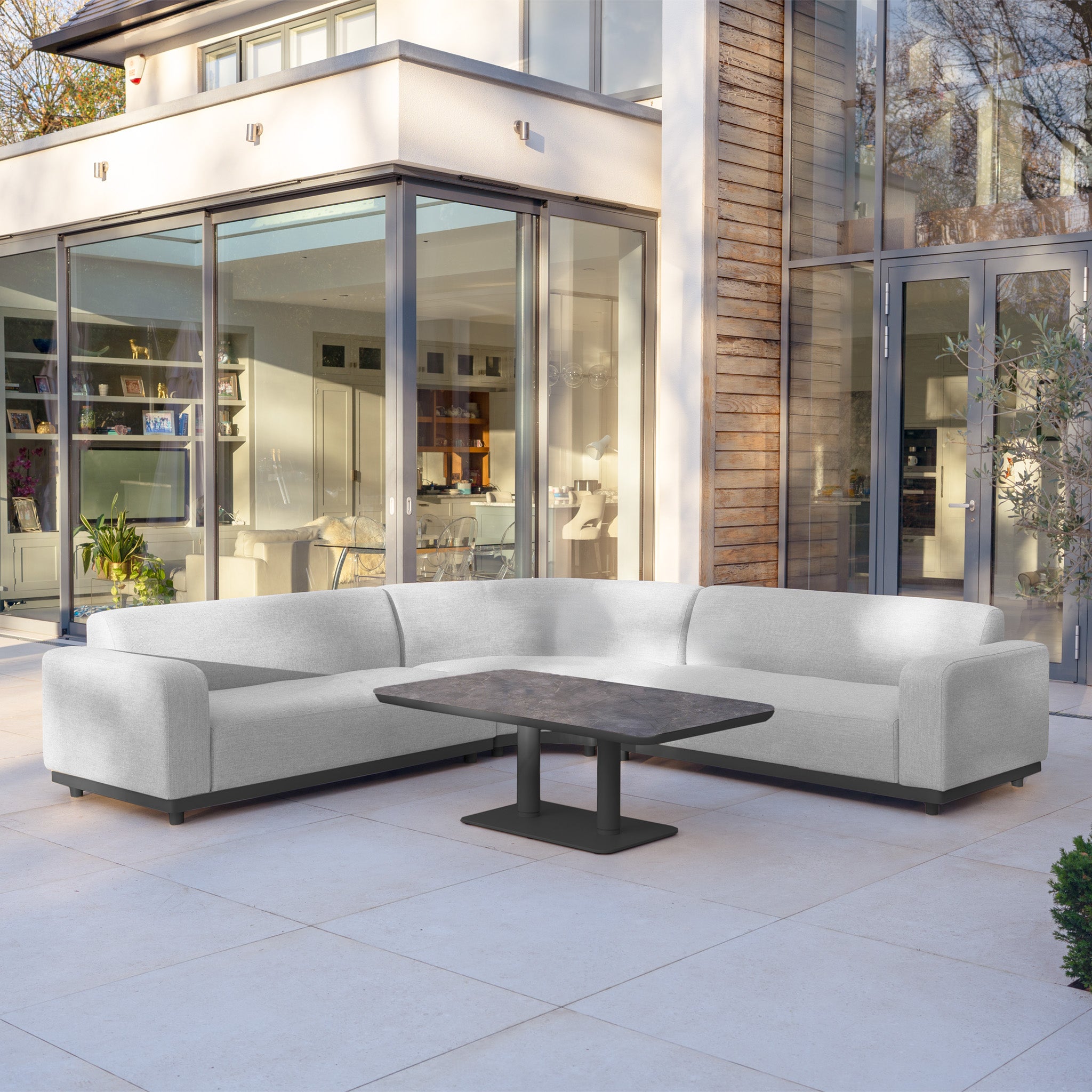 Modern patio featuring the Luna Outdoor Fabric Curved Corner Sofa Set with Altura Rising Table in Oyster Grey beside a glass-walled house, highlighting stylish and durable all-weather garden furniture.