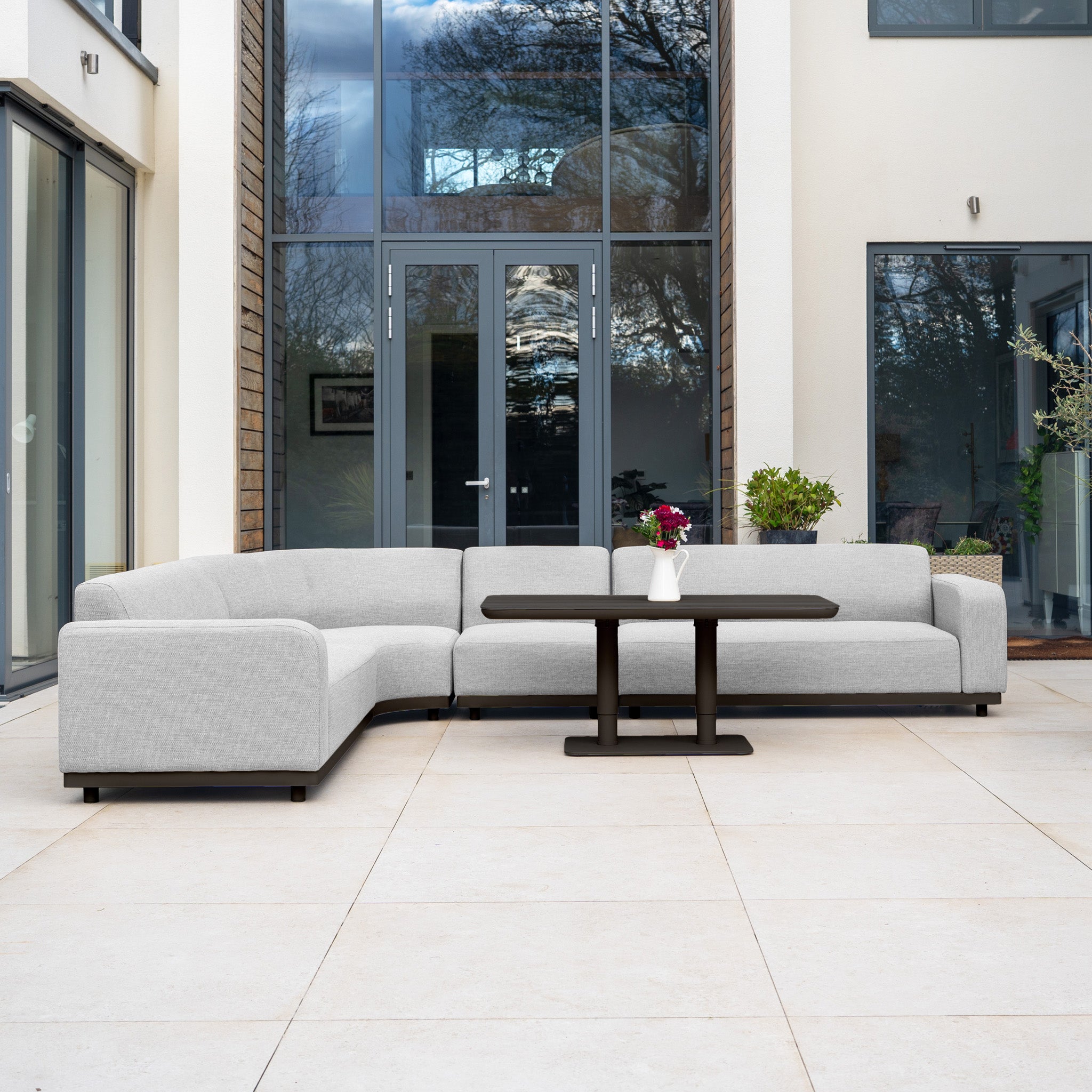Modern outdoor patio featuring the Luna Outdoor Fabric Large Curved Corner Sofa Set in Oyster Grey with an Altura Rising Table, placed by glass doors and accented with potted plants.