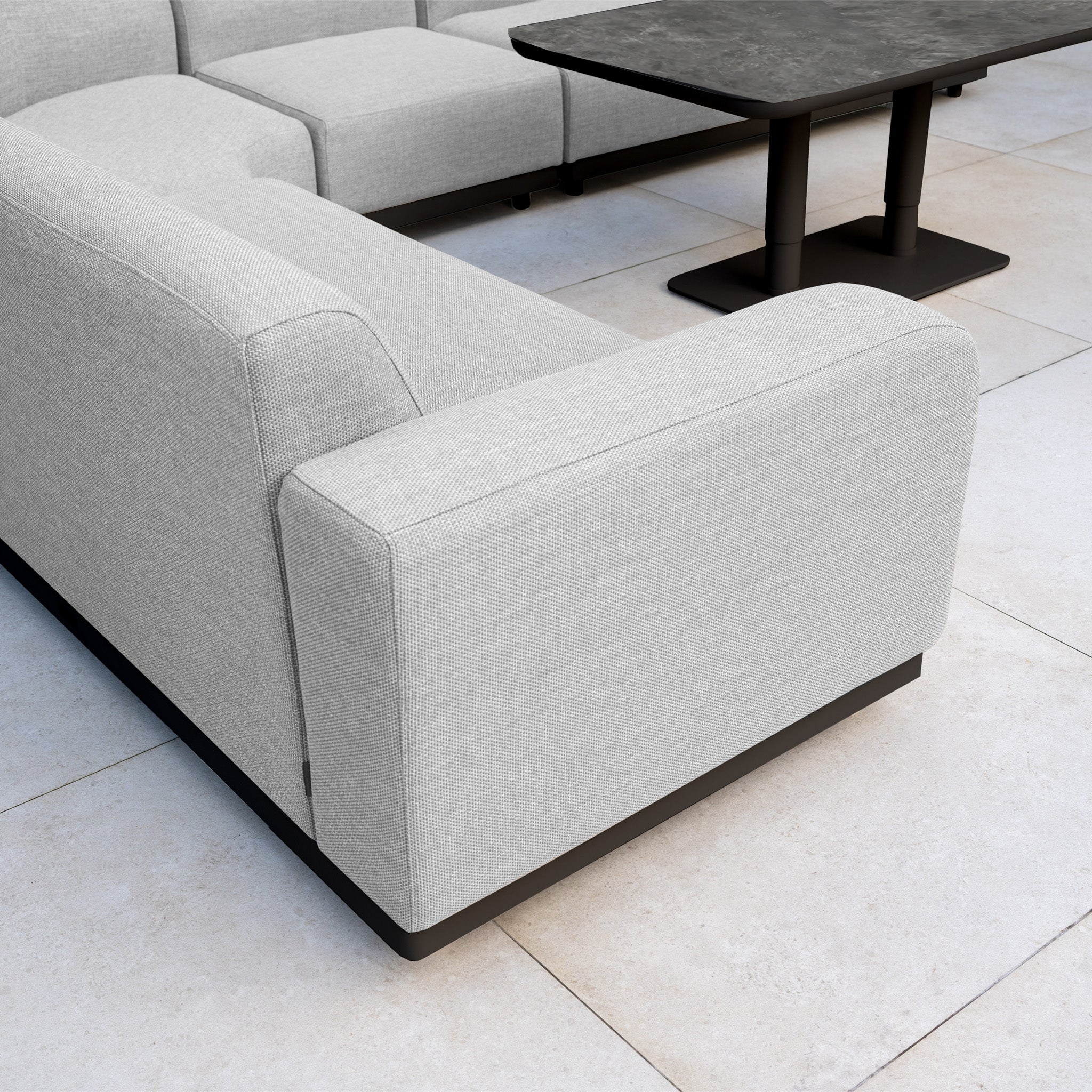 The Luna Outdoor Fabric Large Curved Corner Sofa Set with Altura Rising Table in Oyster Grey features a light grey curved sofa and black rectangular rising table, arranged on a tiled floor.