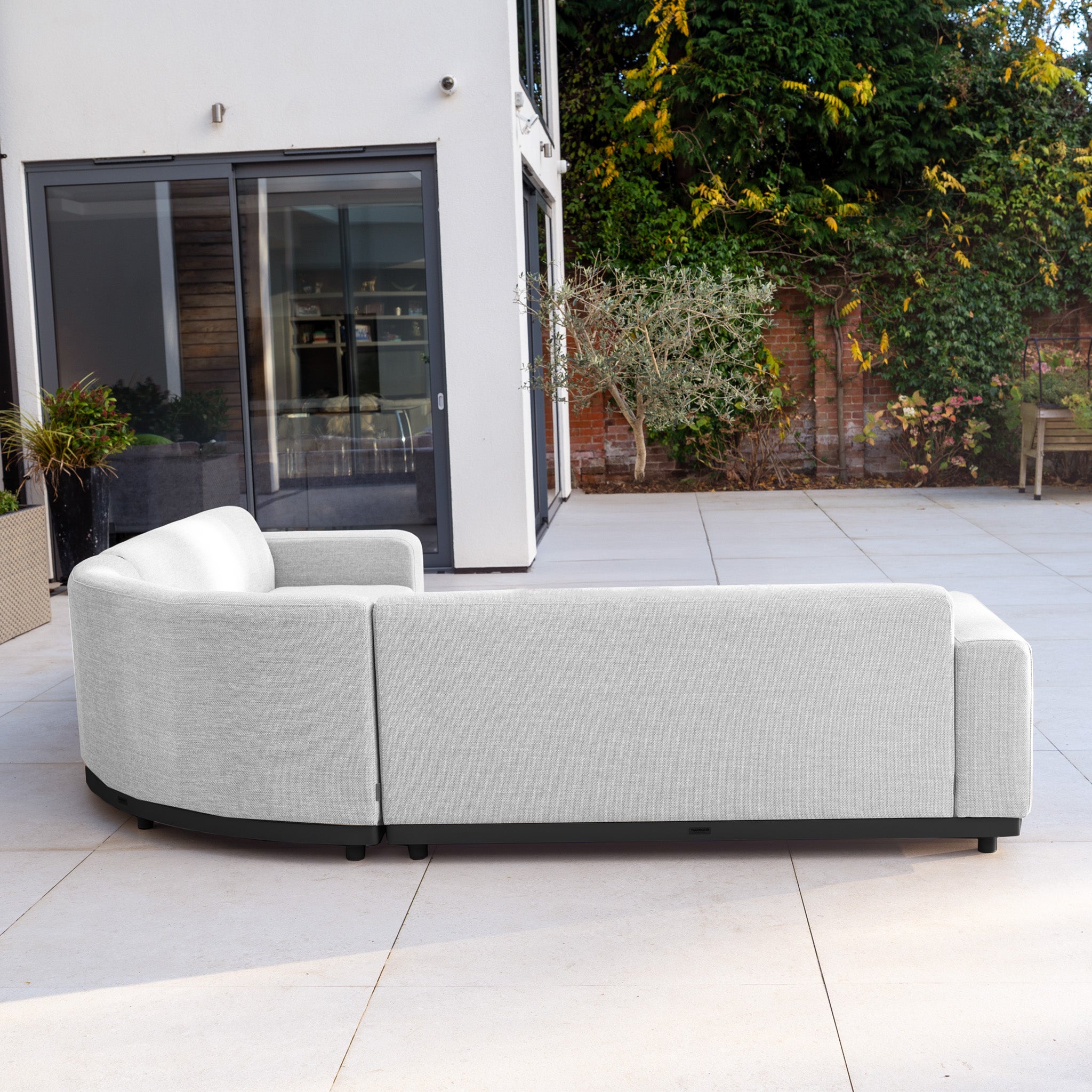 The Luna Outdoor Fabric Curved Corner Sofa Set with Altura Rising Table in Oyster Grey offers stylish, all-weather garden furniture—perfect for relaxing outdoors on a spacious patio by large glass doors.