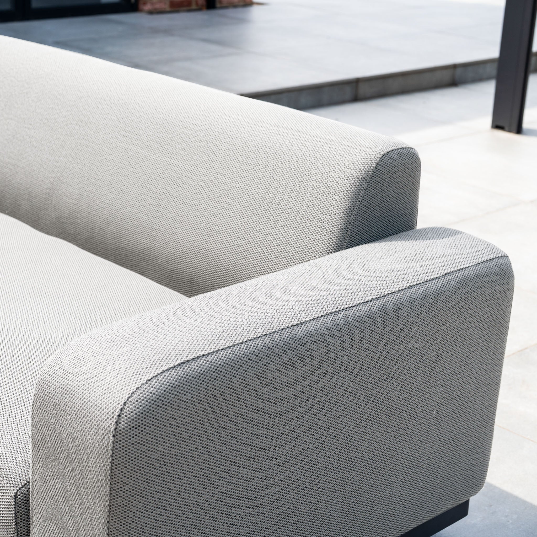 The Luna Outdoor Fabric Large Corner Group Set with Firepit Coffee Table in Oyster Grey sits on a tiled patio in bright sunlight, showcasing its stylish design and durable all-weather fabric.