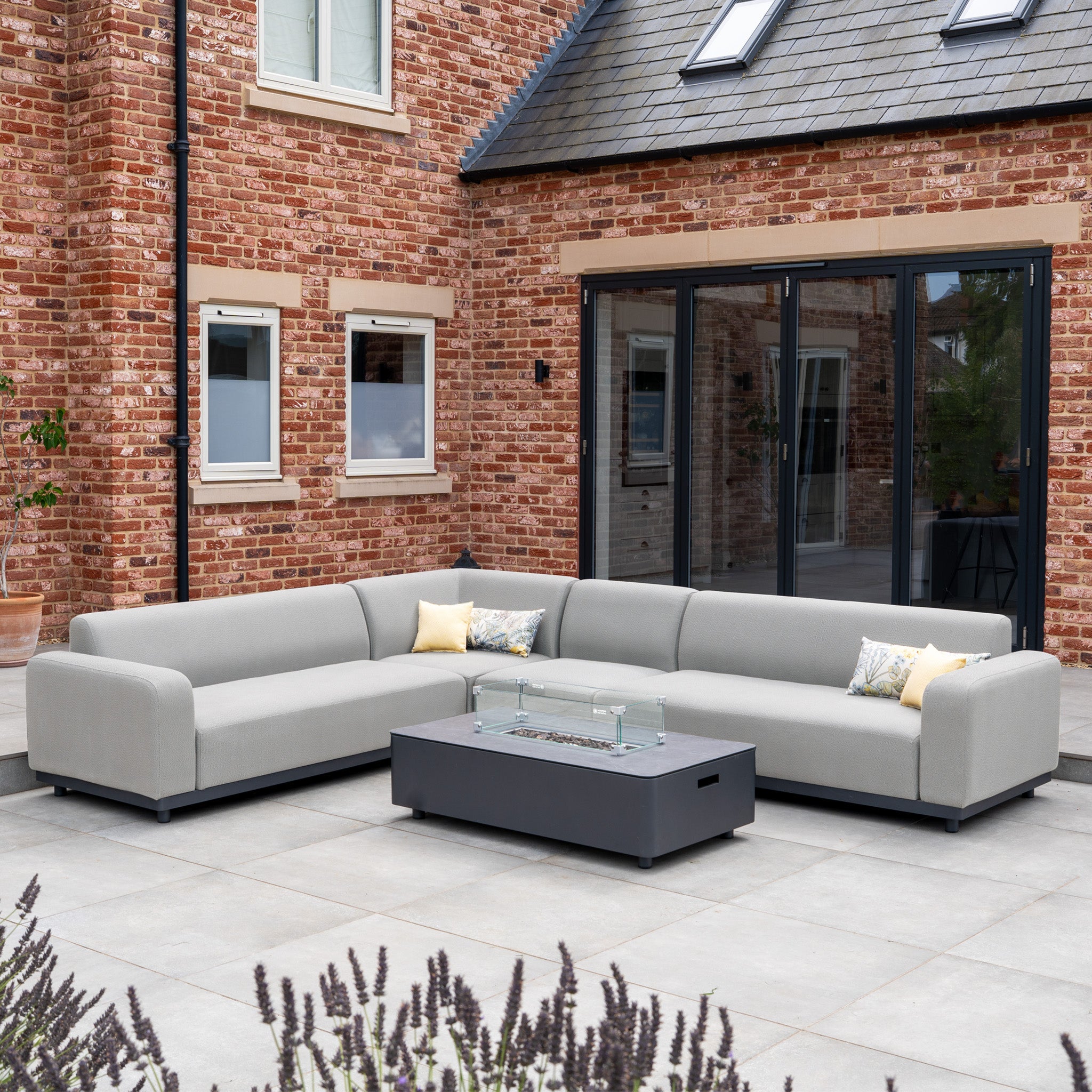 The Luna Outdoor Fabric Large Corner Group Set with Firepit Coffee Table in Oyster Grey sits on a patio beside a brick house.