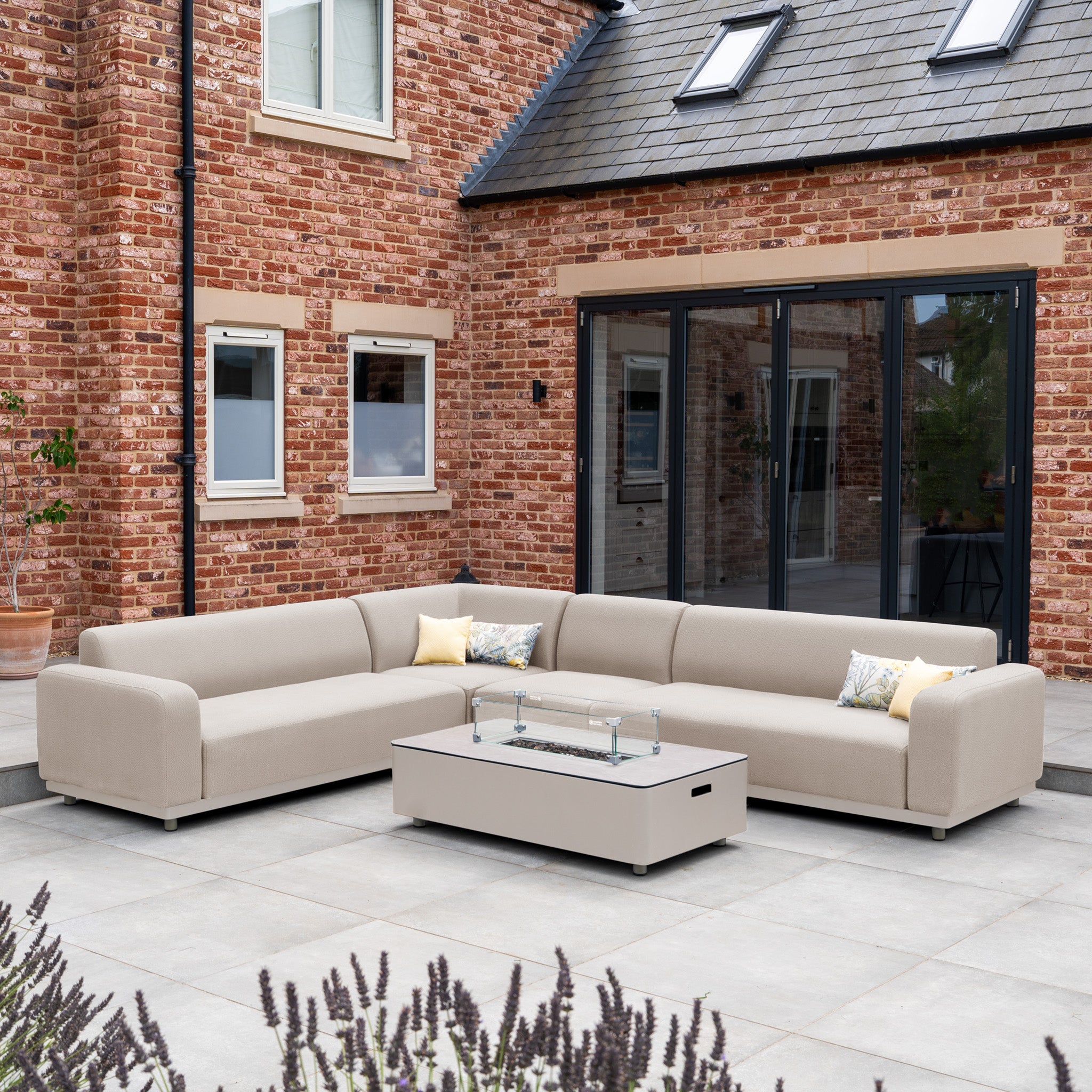 Outdoor patio featuring the Luna Outdoor Fabric Large Corner Group Set with Firepit Coffee Table in Fawn, set against a brick house backdrop.