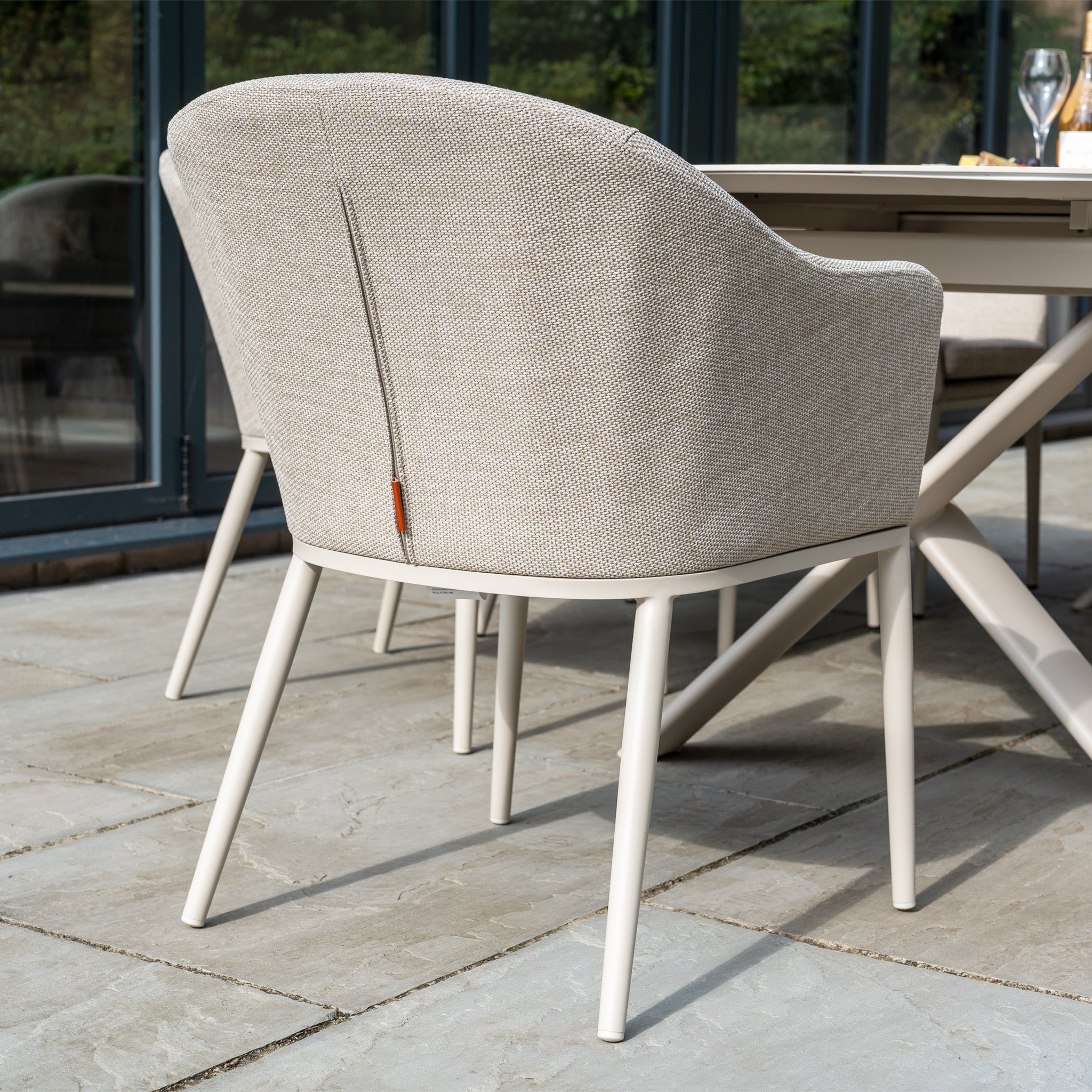 The Luna 8 Seat Outdoor Fabric Oval Dining Set with Albatross Extending Table in Fawn, featuring UV-resistant cushions, sits on a stone patio near glass doors.