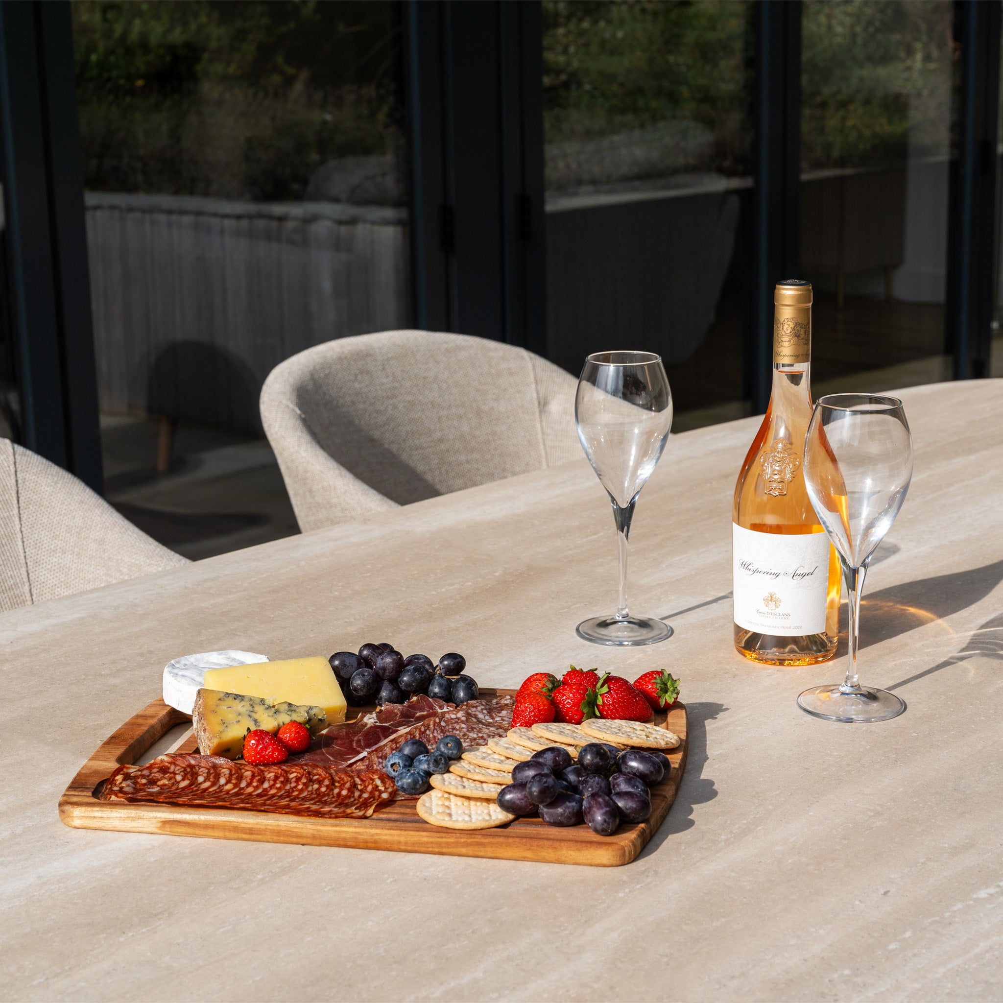 A charcuterie board with cheese, fruit, and meat sits beside wine glasses and a wine bottle on a patio table, part of the Luna 8 Seat Outdoor Fabric Oval Ceramic Dining Set in Fawn, designed for all-weather durability.