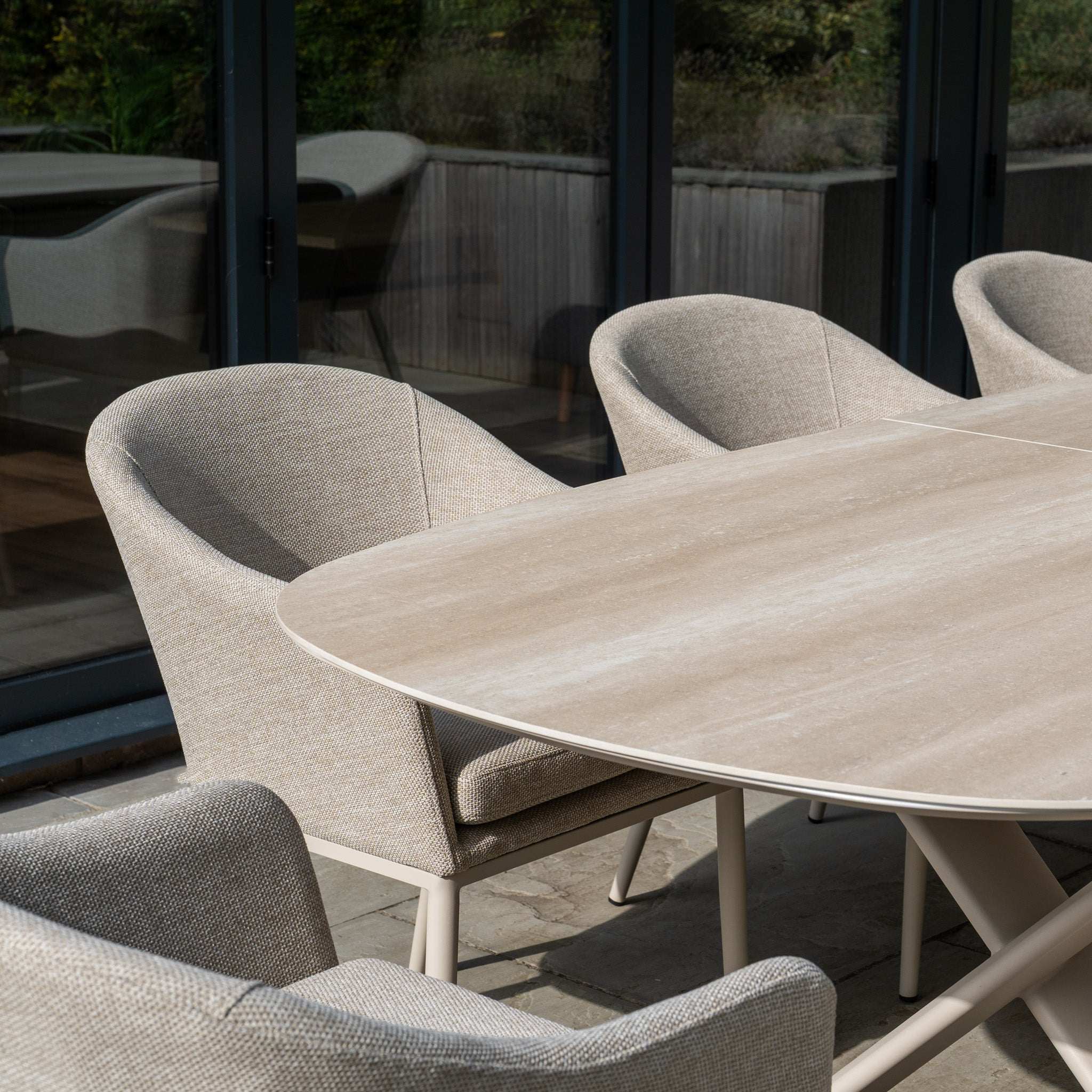 The Luna 8 Seat Outdoor Fabric Oval Dining Set with Albatross Extending Table in Fawn features UV resistant light beige cushions on chairs around a wooden table, perfect for patios near glass doors.