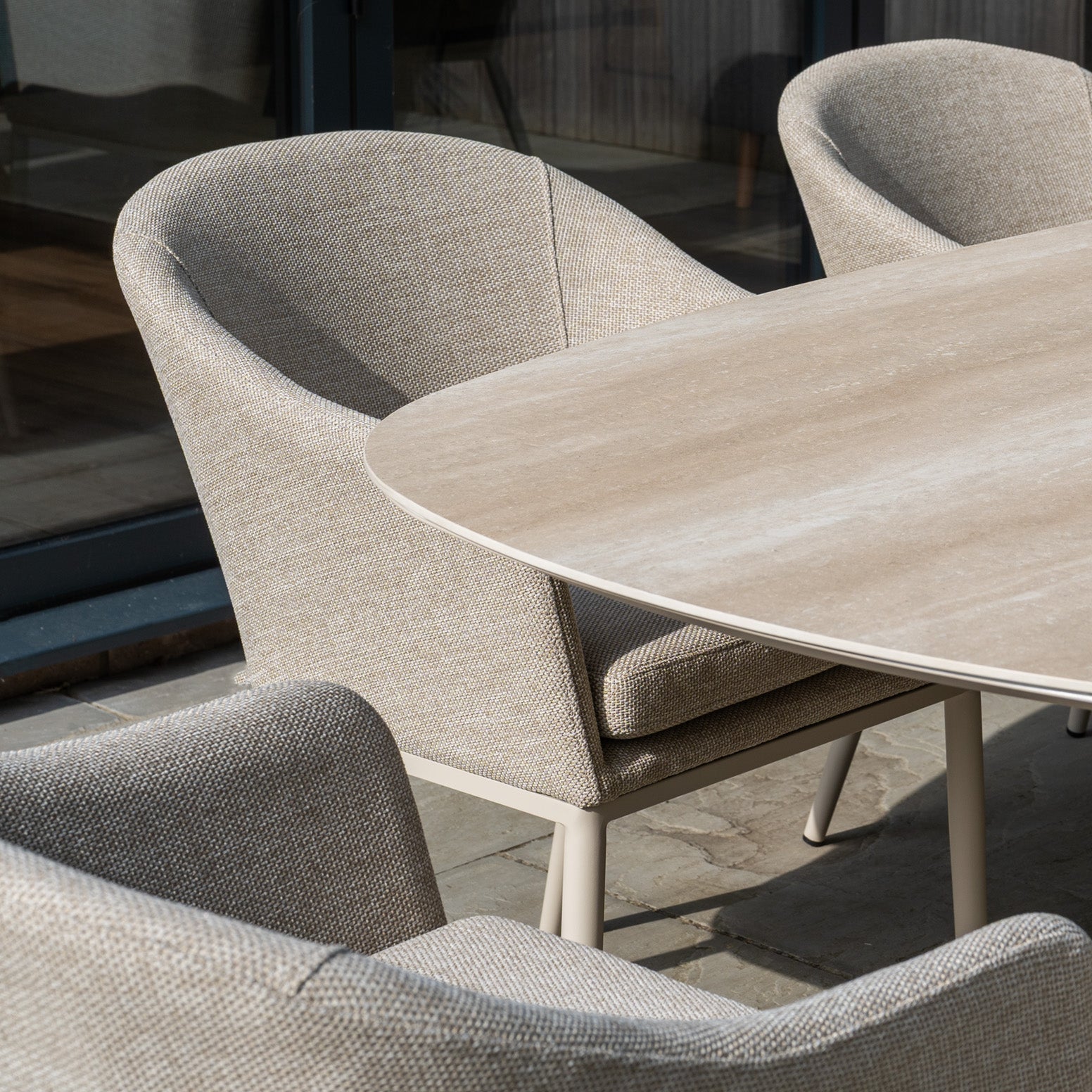 The Luna 8 Seat Outdoor Fabric Oval Ceramic Dining Set in Fawn features upholstered chairs and a ceramic oval table on a stone patio by glass doors.
