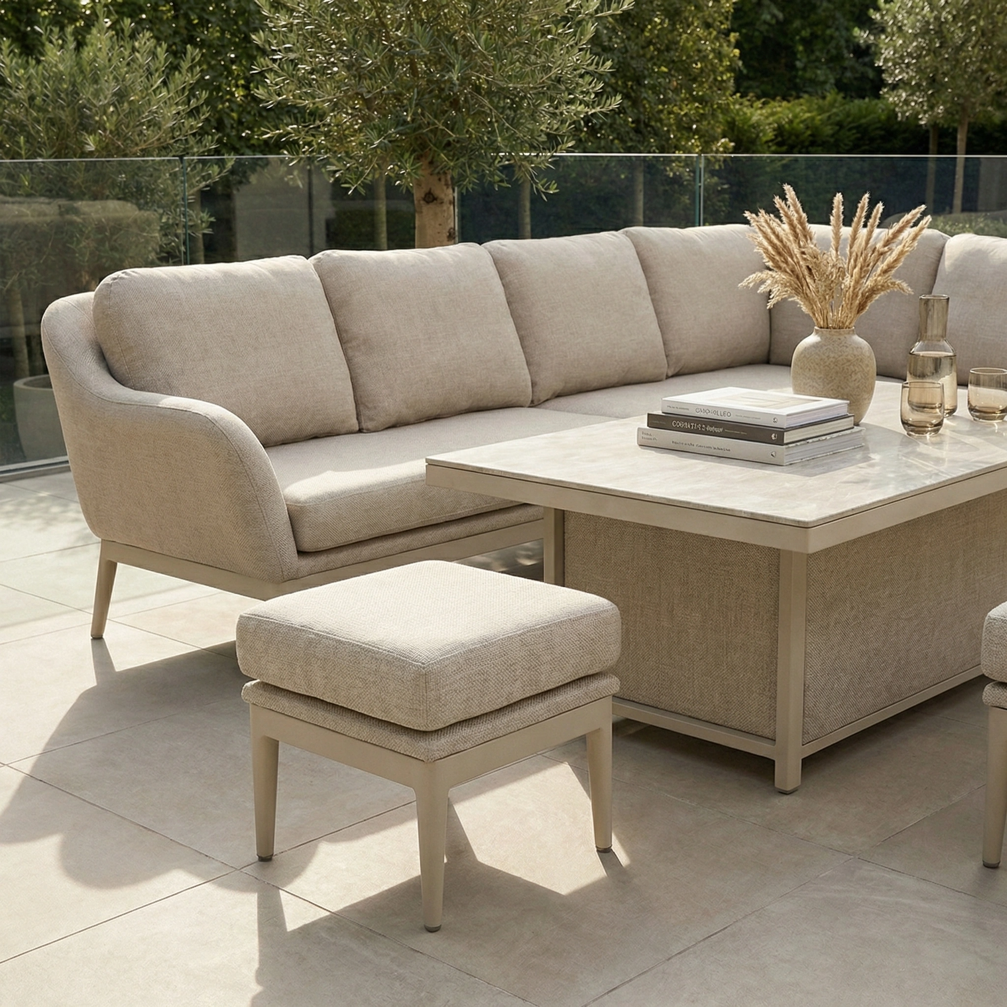 The Luna Outdoor Fabric Rectangular Corner Dining Set with Rising Table in Fawn (Right Hand) sits on a tiled patio, styled with books and a vase, offering an inviting all-weather setup complete with an ottoman.