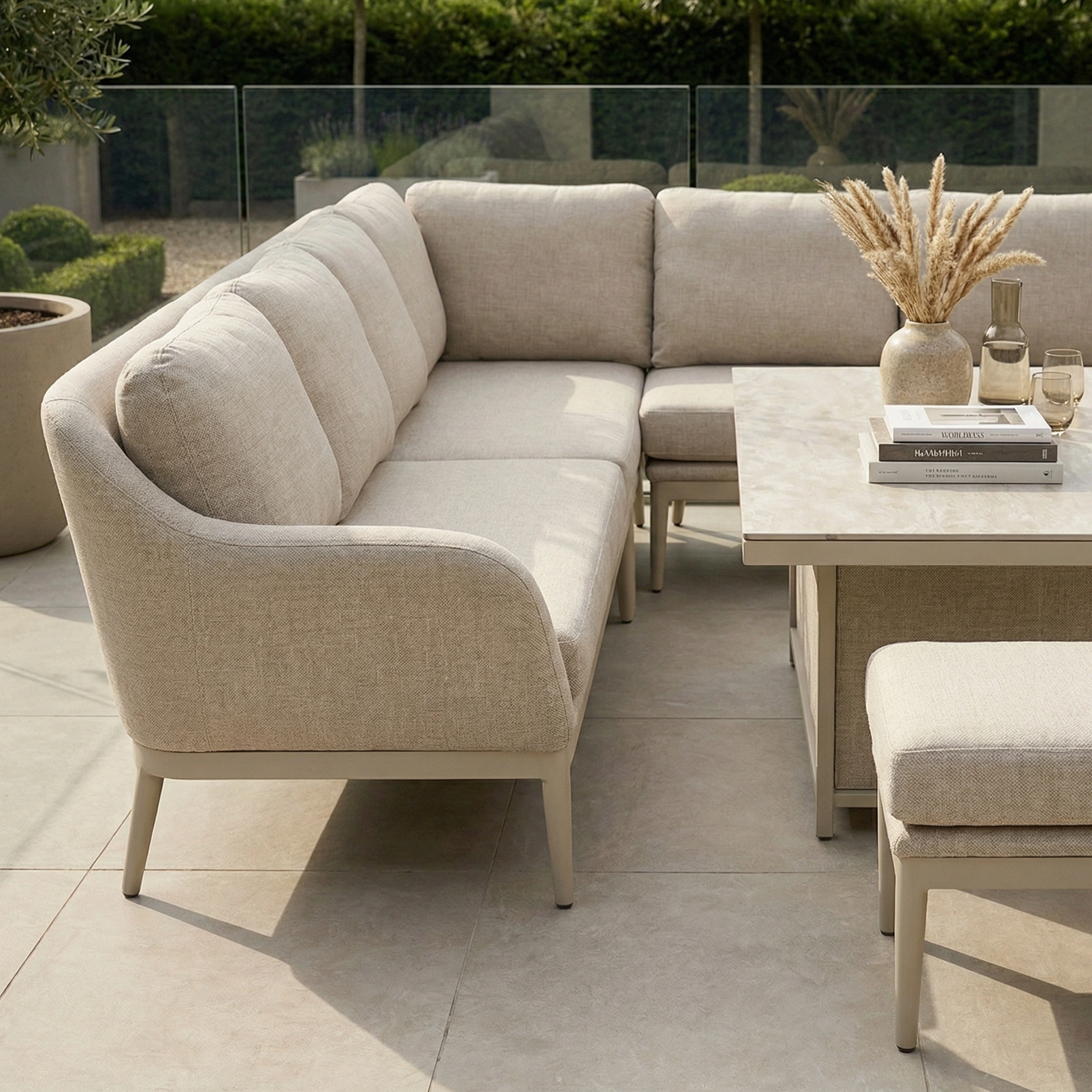 The Luna Outdoor Fabric Rectangular Corner Dining Set with Rising Table in Fawn (Right Hand) is arranged on a tiled patio, styled with books, vases, and pampas grass.