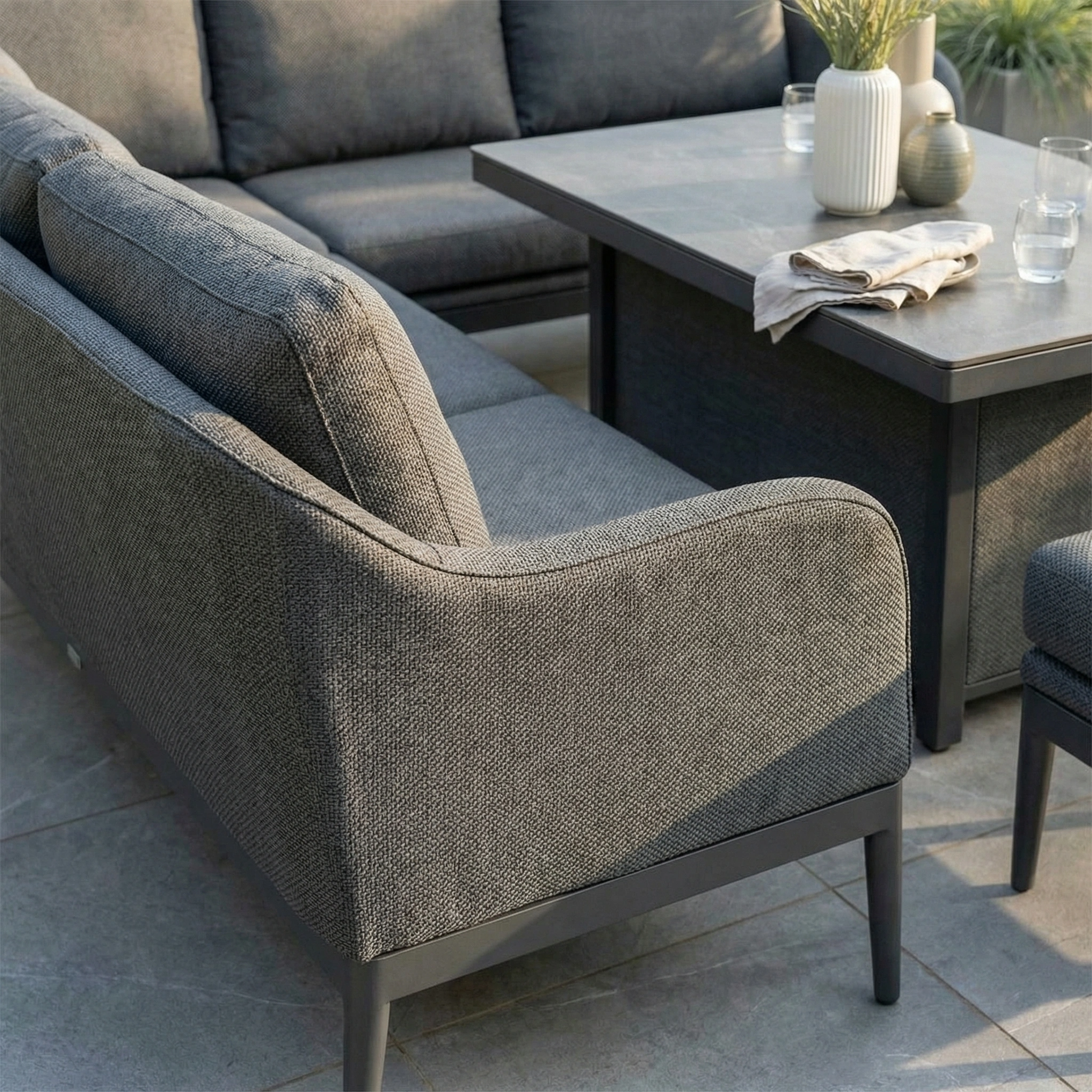 The Luna Outdoor Fabric Rectangular Corner Dining Set with Rising Table in Grey (Right Hand) features durable all-weather materials, decorative vases, glasses, and a napkin on a tiled patio for lasting outdoor style.