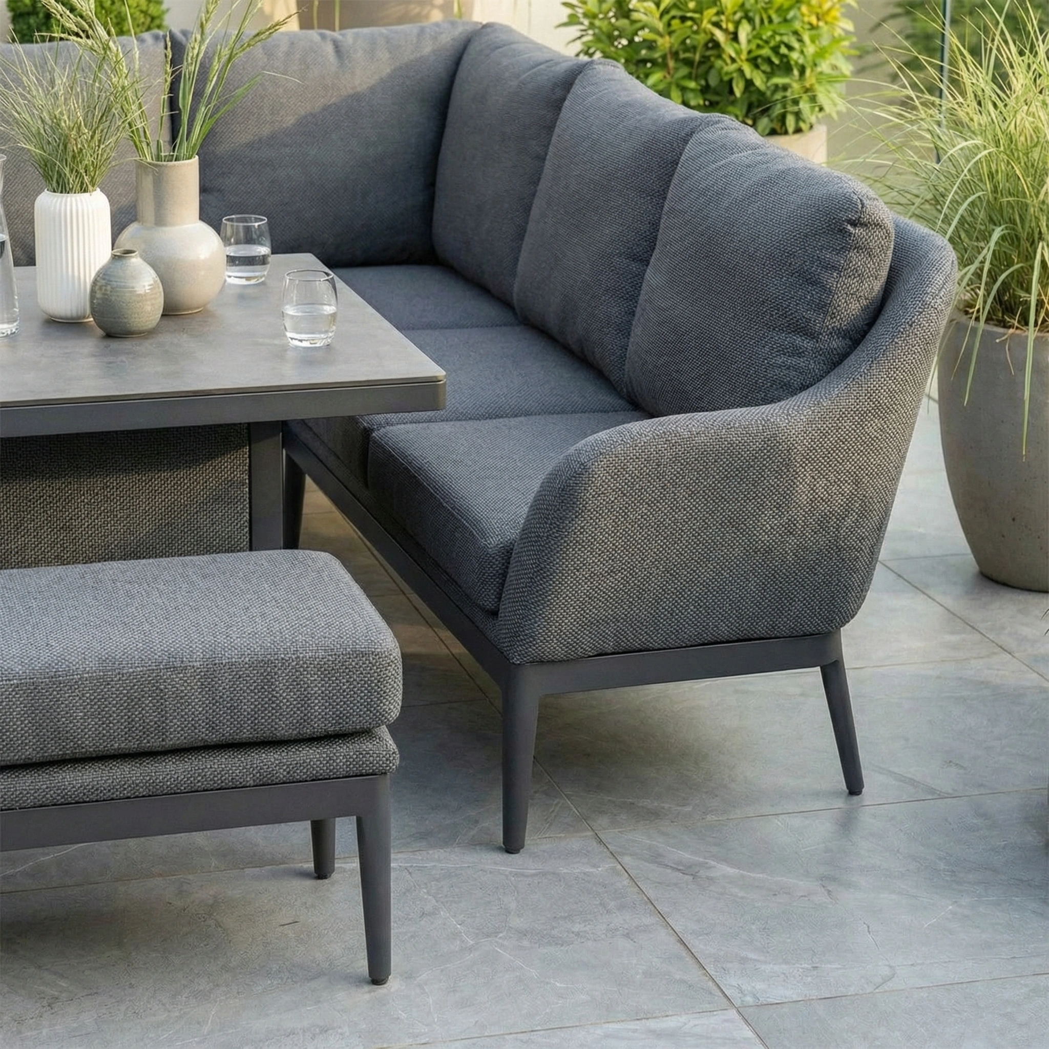 The Luna Outdoor Fabric Rectangular Corner Dining Set with Rising Table in Grey (Right Hand) sits on a tiled patio, featuring a weather-resistant sectional and bench, potted plants, and a decor-accented table.