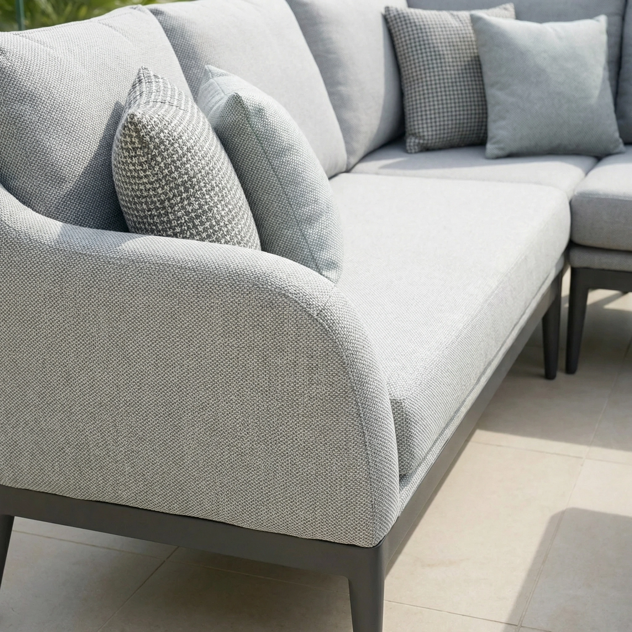 A light grey outdoor sofa with matching cushions sits on a tiled patio in bright natural light, perfectly complementing the Luna Outdoor Fabric Rectangular Corner Dining Set with Rising Table in Oyster Grey (Right Hand).