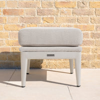 The Luna Stool in Fawn features quick-dry foam cushions and four legs, shown here against a yellow brick wall on a tiled floor—an all-weather outdoor stool ideal for any stylish space.