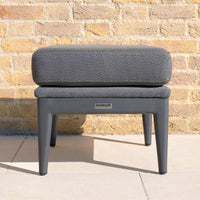 The Luna Stool in Grey, labeled "Harbour" and designed for all-weather outdoor use, sits on a tiled floor against a yellow brick wall.