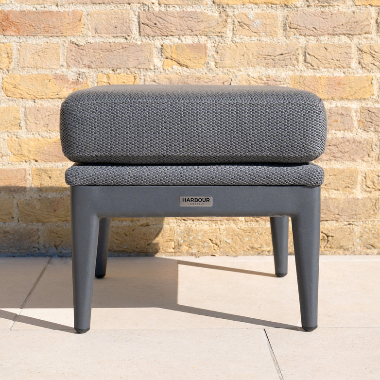 The Luna Stool in Grey, labeled "Harbour" and designed for all-weather outdoor use, sits on a tiled floor against a yellow brick wall.