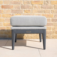The Luna Stool in Oyster Grey features quick-dry cushions and sturdy metal legs, making it ideal for all-weather outdoor spaces, as it sits stylishly against a light brick wall in the sunlight.