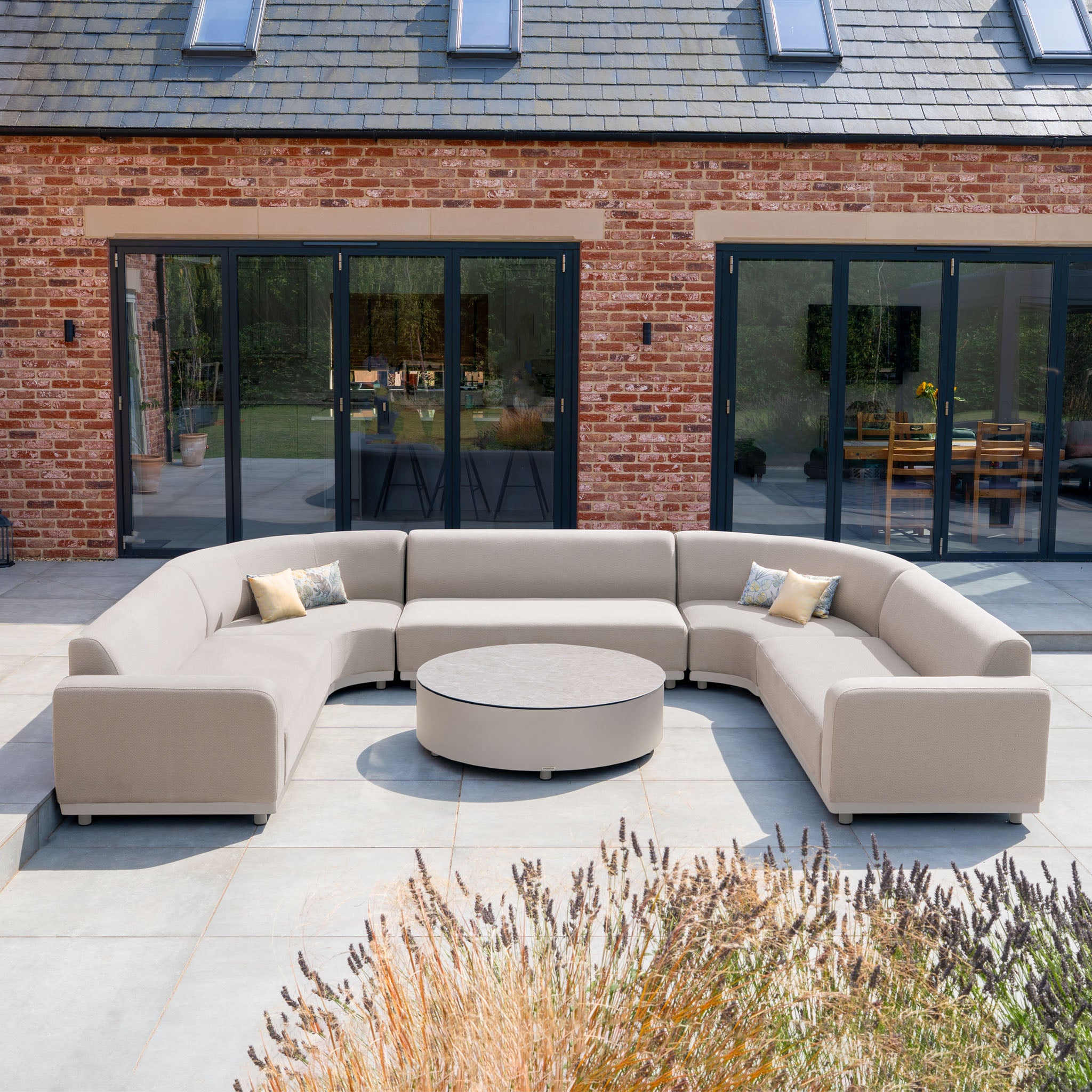 The Luna U-Shape Outdoor Fabric Curved Corner Group with Round Coffee Table in Fawn sits on a patio before a brick house with glass doors, crafted from durable all-weather materials for lasting outdoor elegance.