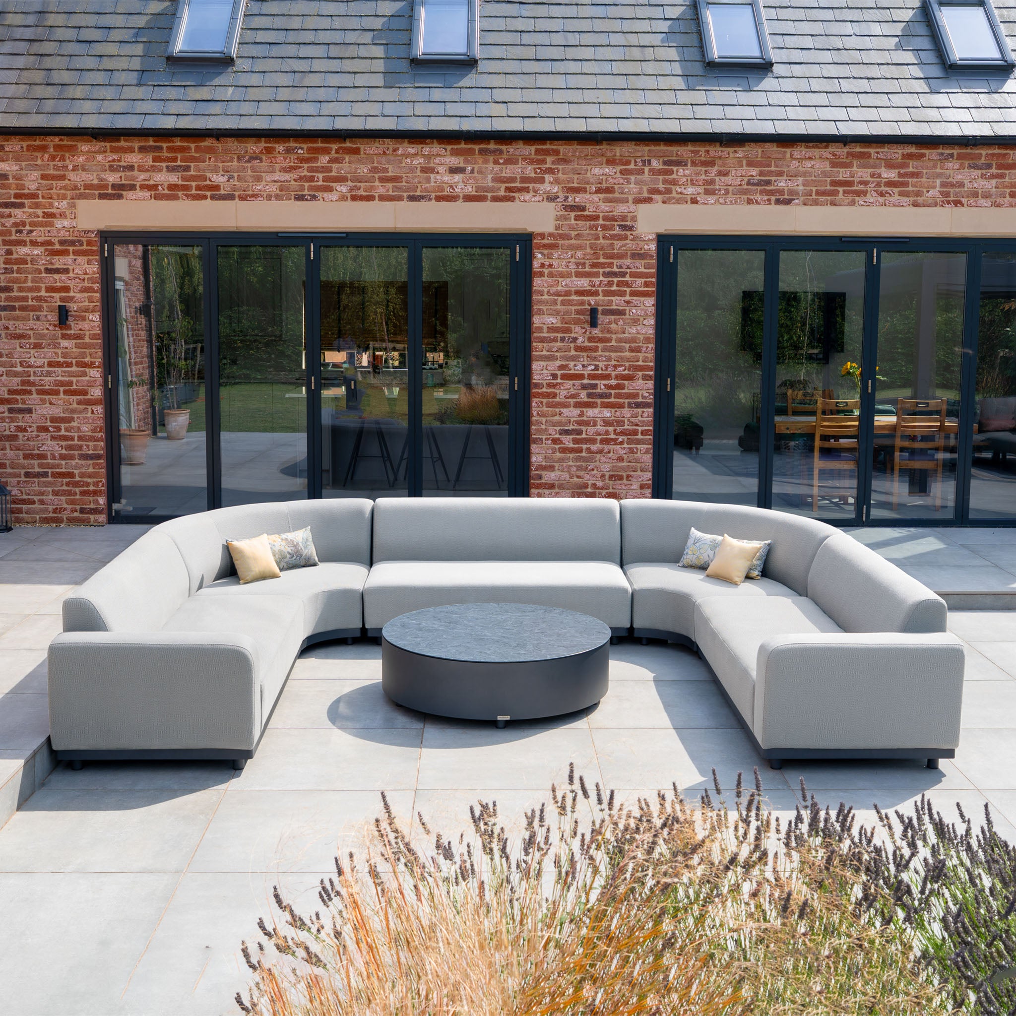 The Luna U-Shape Outdoor Fabric Curved Corner Group with Round Coffee Table in Oyster Grey sits on a patio by a brick house with sliding glass doors, featuring an aluminium frame for stylish, all-weather durability.