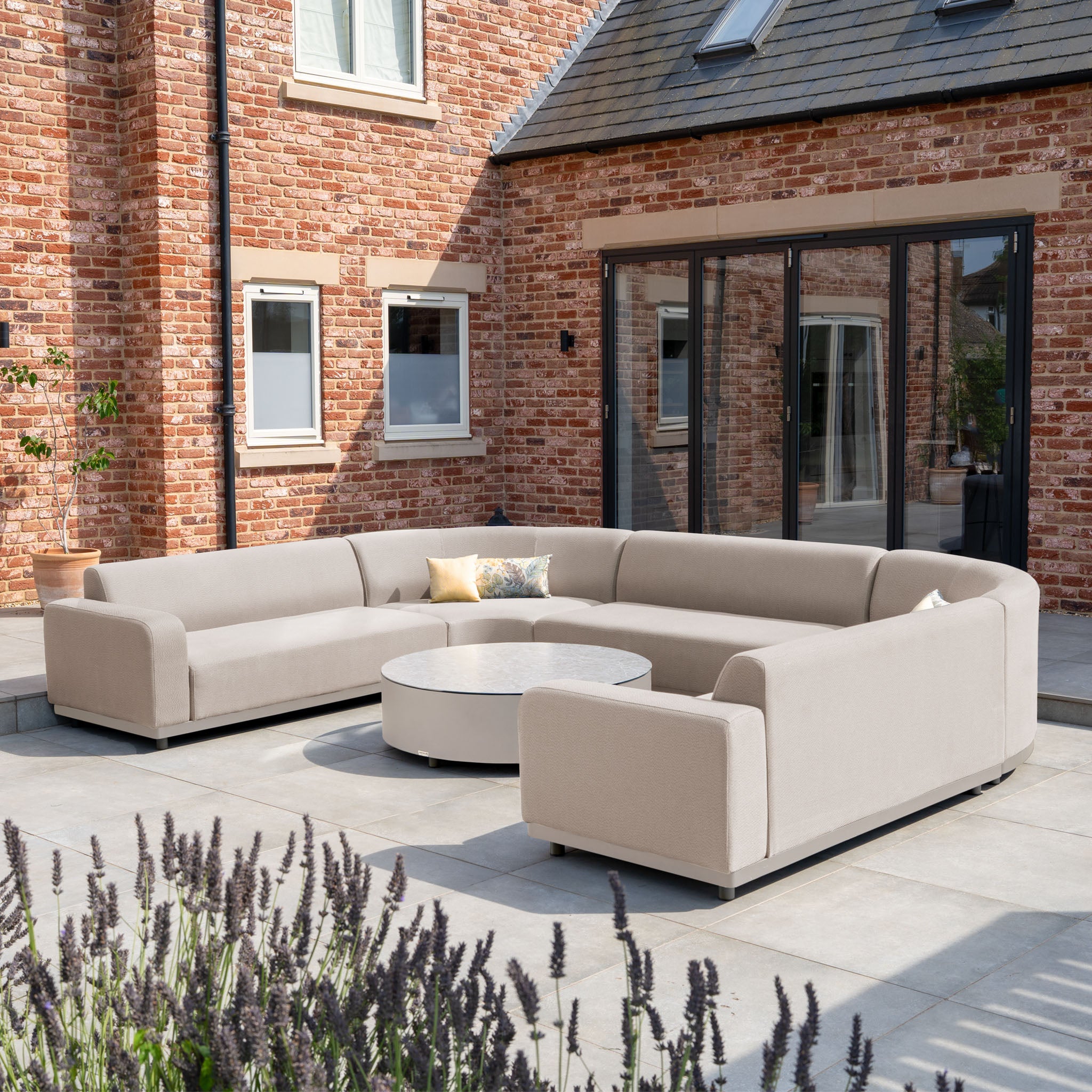 The Luna U-Shape Outdoor Fabric Curved Corner Group with Round Coffee Table in Fawn is stylish all-weather patio furniture, perfect for relaxing outdoors in front of a brick house.