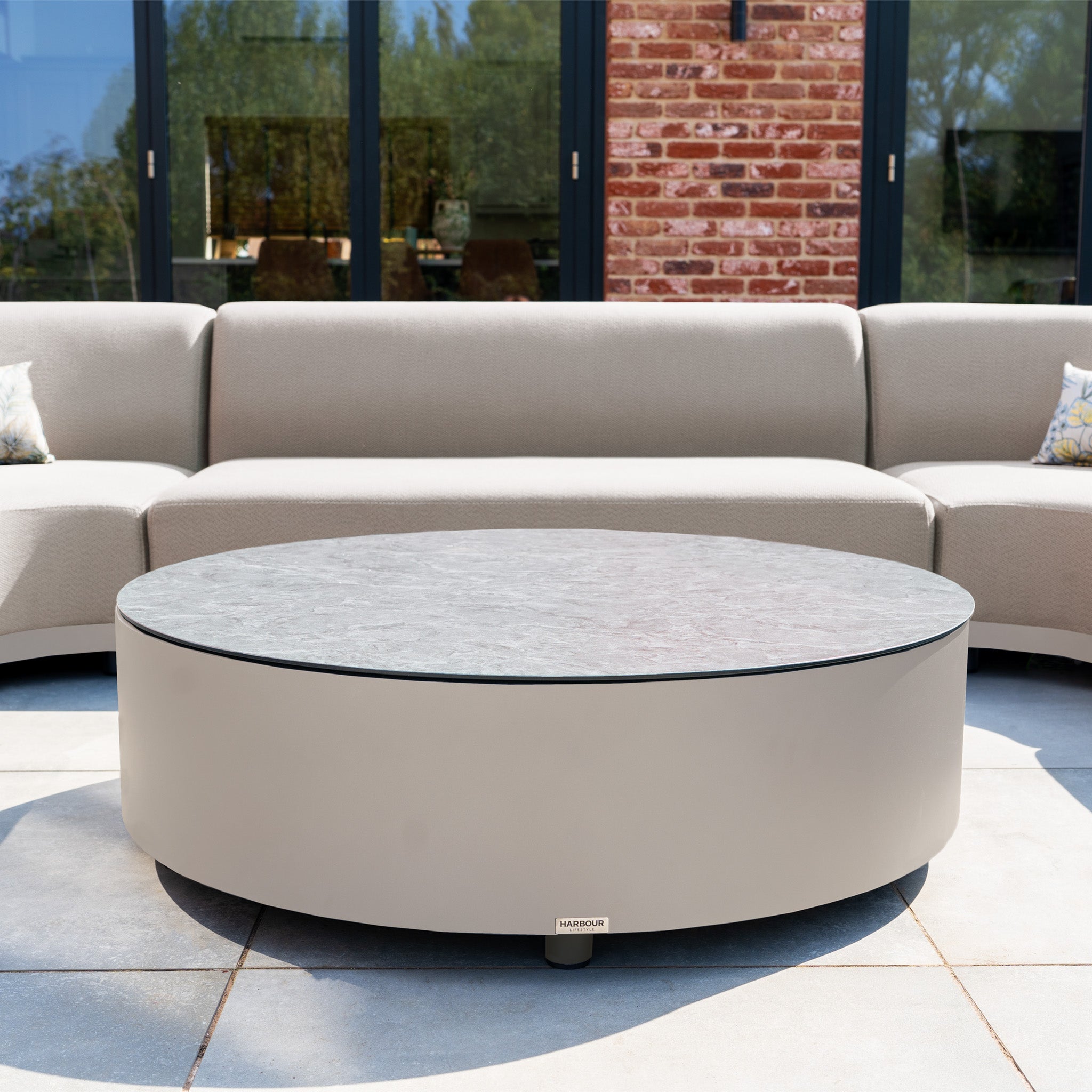 The Luna U-Shape Outdoor Fabric Curved Corner Group in Fawn, with a round coffee table, is set on a patio alongside beige sofas and glass doors in the background.
