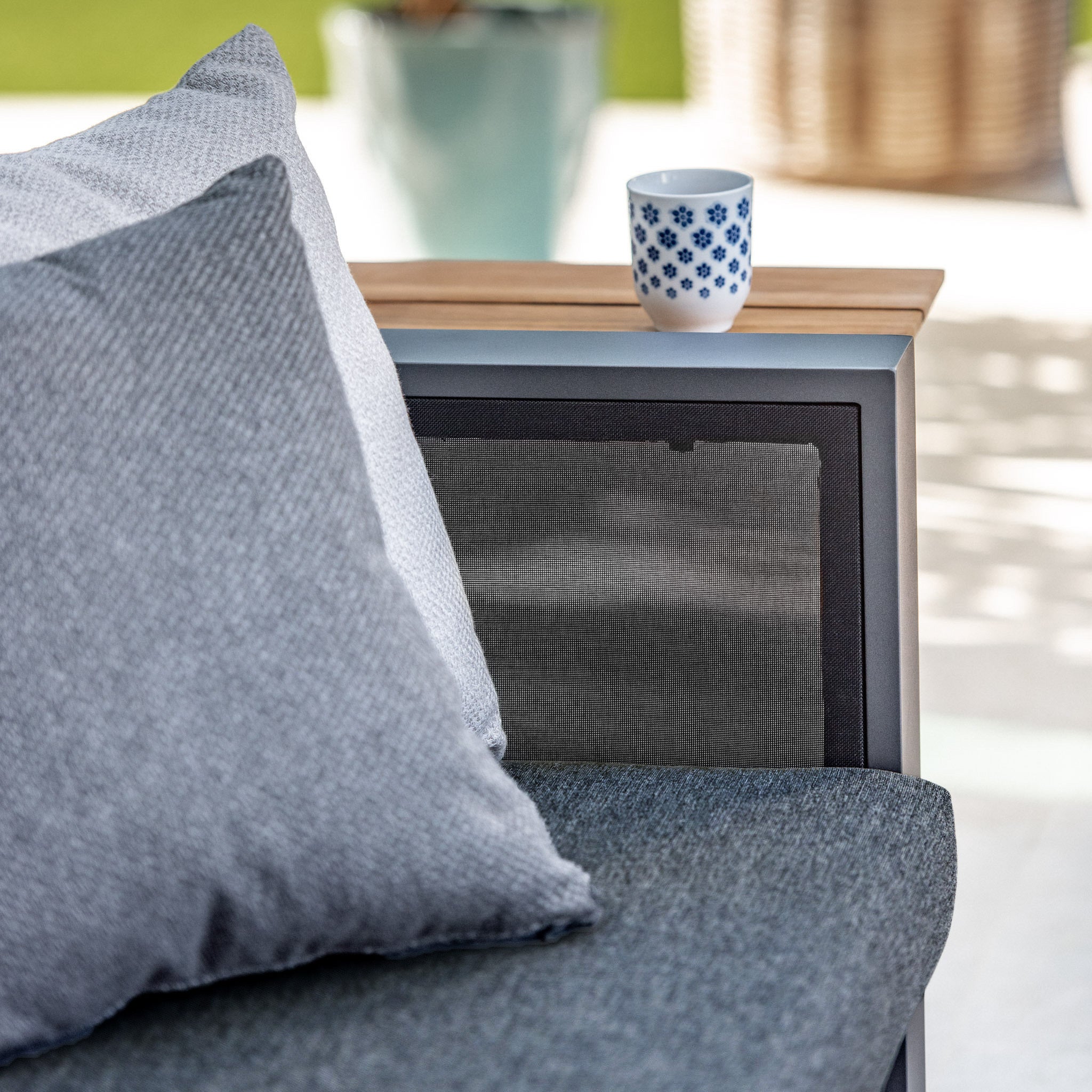 The Manhattan Armchair with Teak Gas Lift Arm in Charcoal is perfect for stylish outdoor arrangements—pair it with Olefin seat cushions, accent pillows, a wooden side table, and a white mug with blue patterns for a chic patio look.
