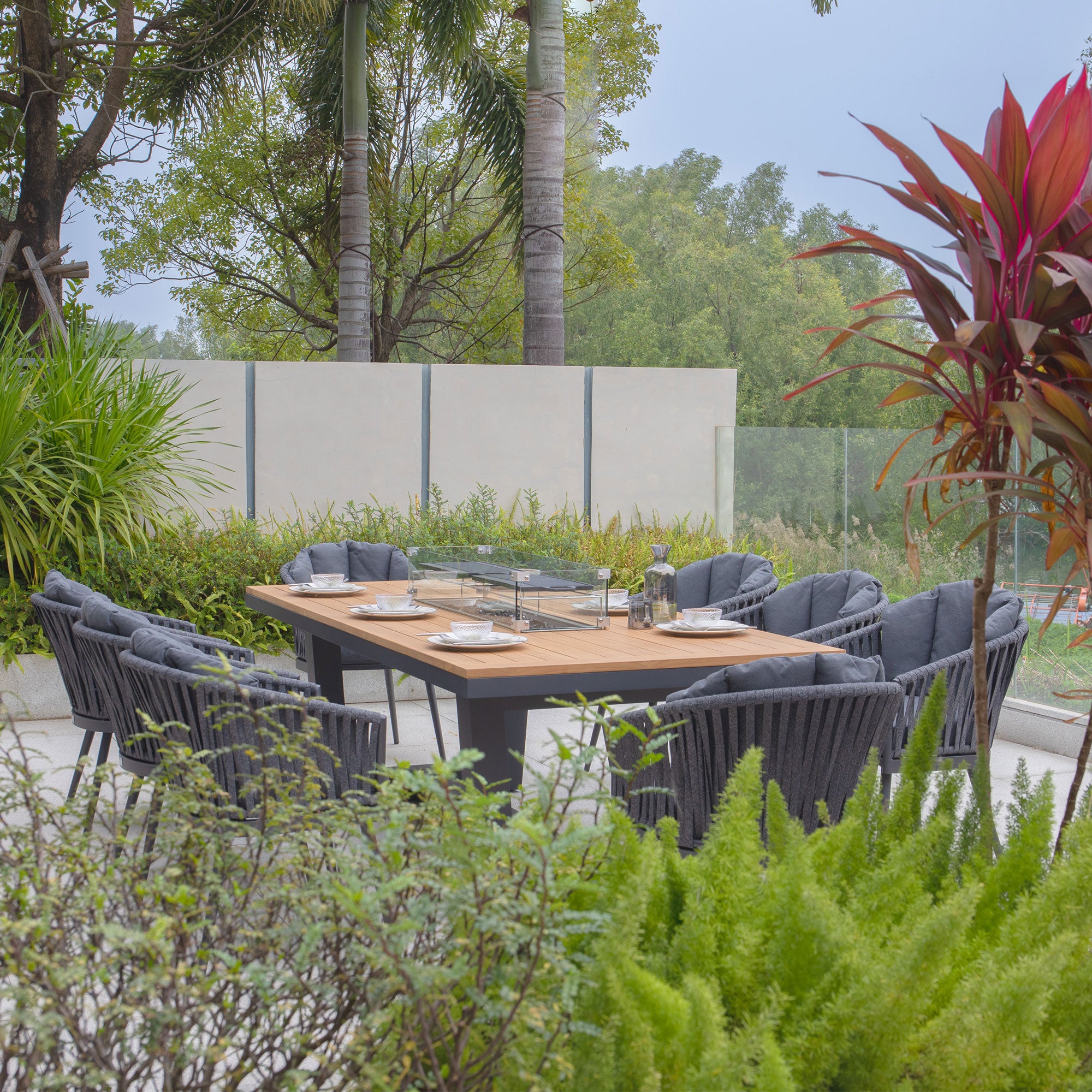 Outdoor dining table with eight chairs, featuring the Monterrey 8 Seat Rope Rectangular Teak Firepit Dining Set in Grey, set in a garden surrounded by lush greenery and decorative plants.