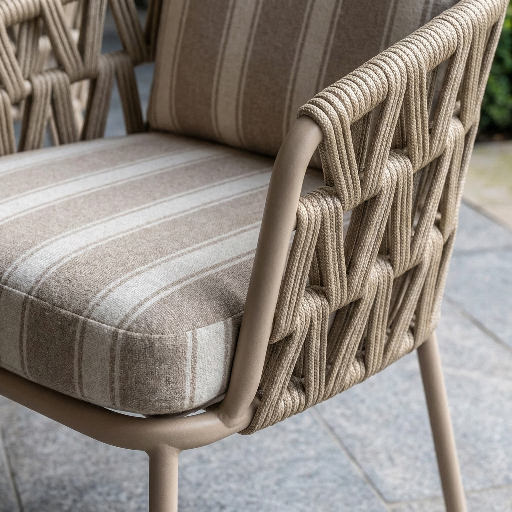Close-up of a modern outdoor chair with woven rope sides and striped beige cushion, part of the Madeira 6 Seat Curved Rectangular Dining Set in Striped Latte, featuring a powder-coated aluminium table on a stone patio.