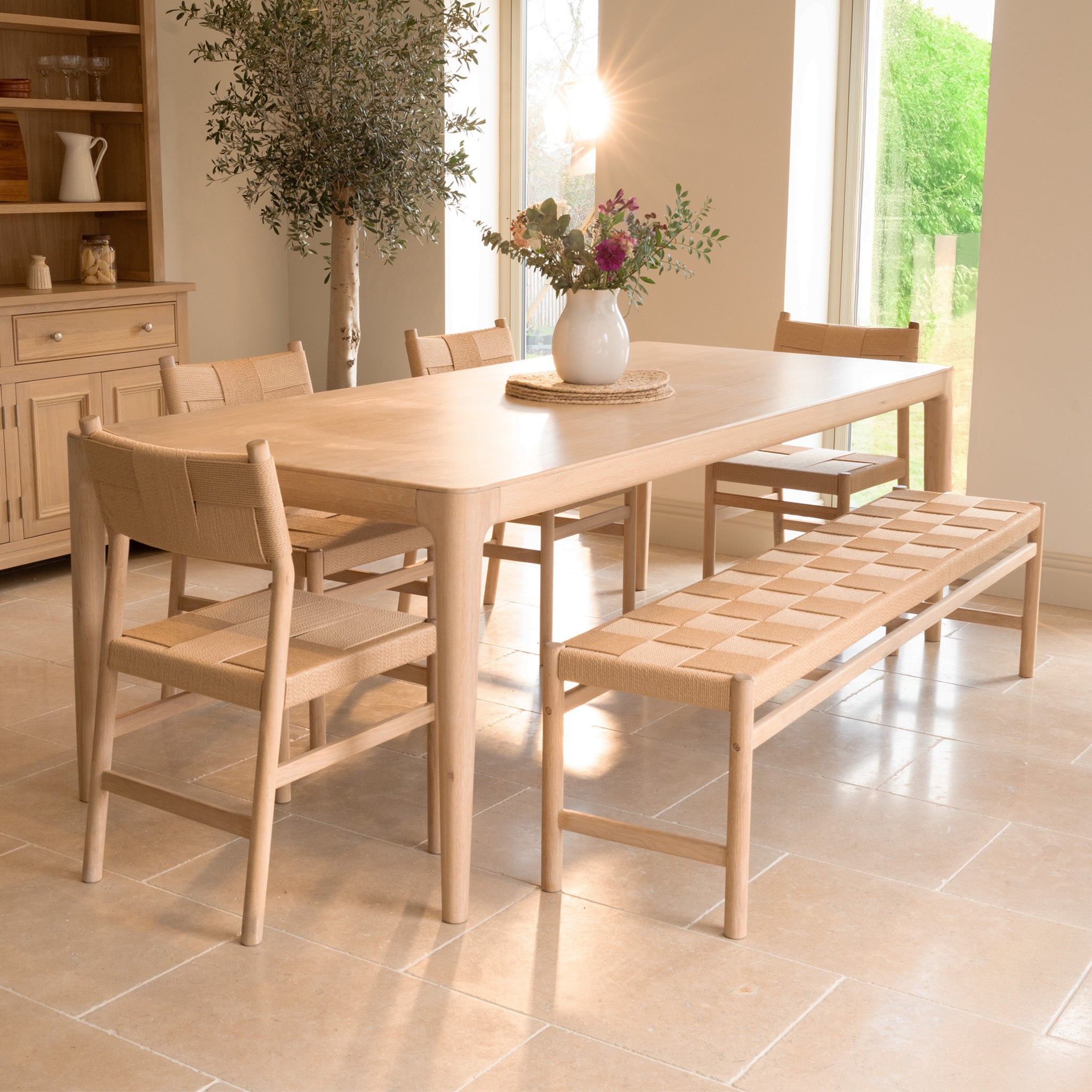 The Marlow 8 Seat Dining Set with Woven Bench in Light Oak, arranged with a vase of flowers, brings style and brightness to any airy dining room.
