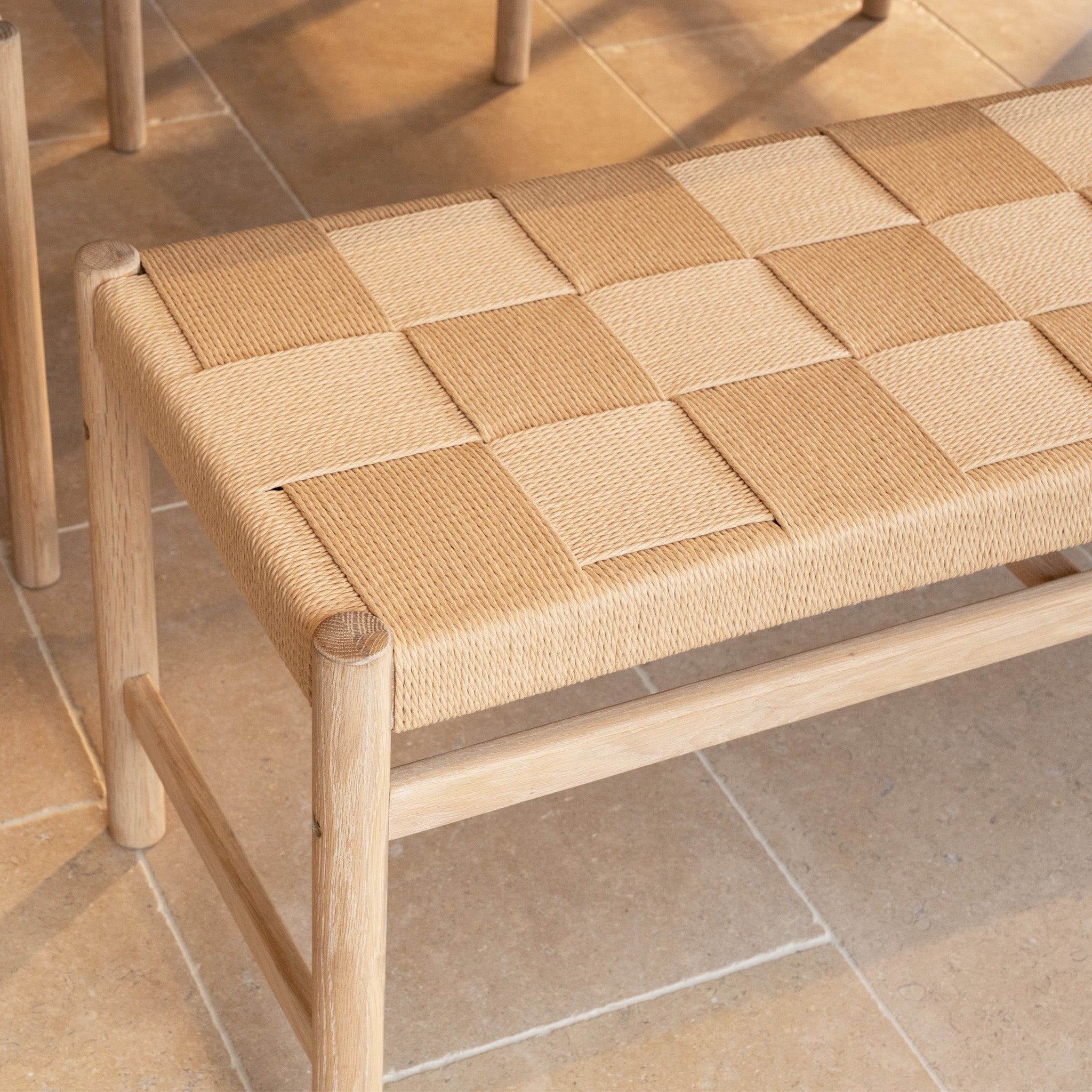 A woven bench with a checkered seat sits on tiled flooring, perfectly complementing the Marlow 8 Seat Dining Set with Woven Bench in Light Oak.