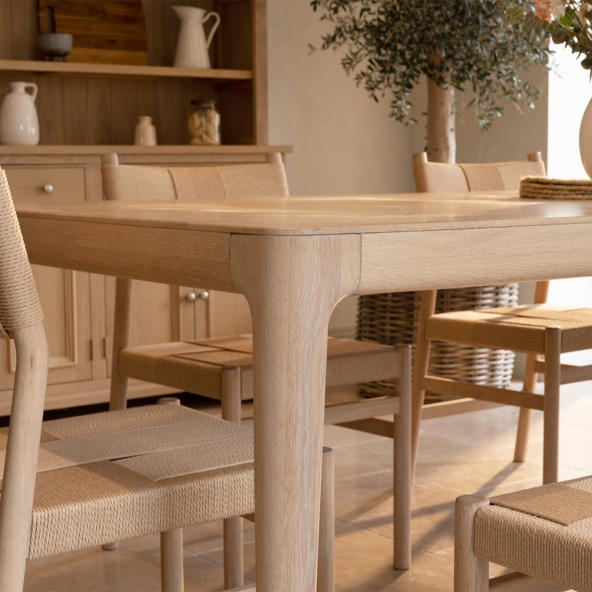 The Marlow 8 Seat Dining Set in Light Oak features a solid oak dining table paired with handwoven dining chairs, beautifully arranged in a sunlit room with a plant and cabinet in the background—perfectly blending style and comfort for your gatherings.